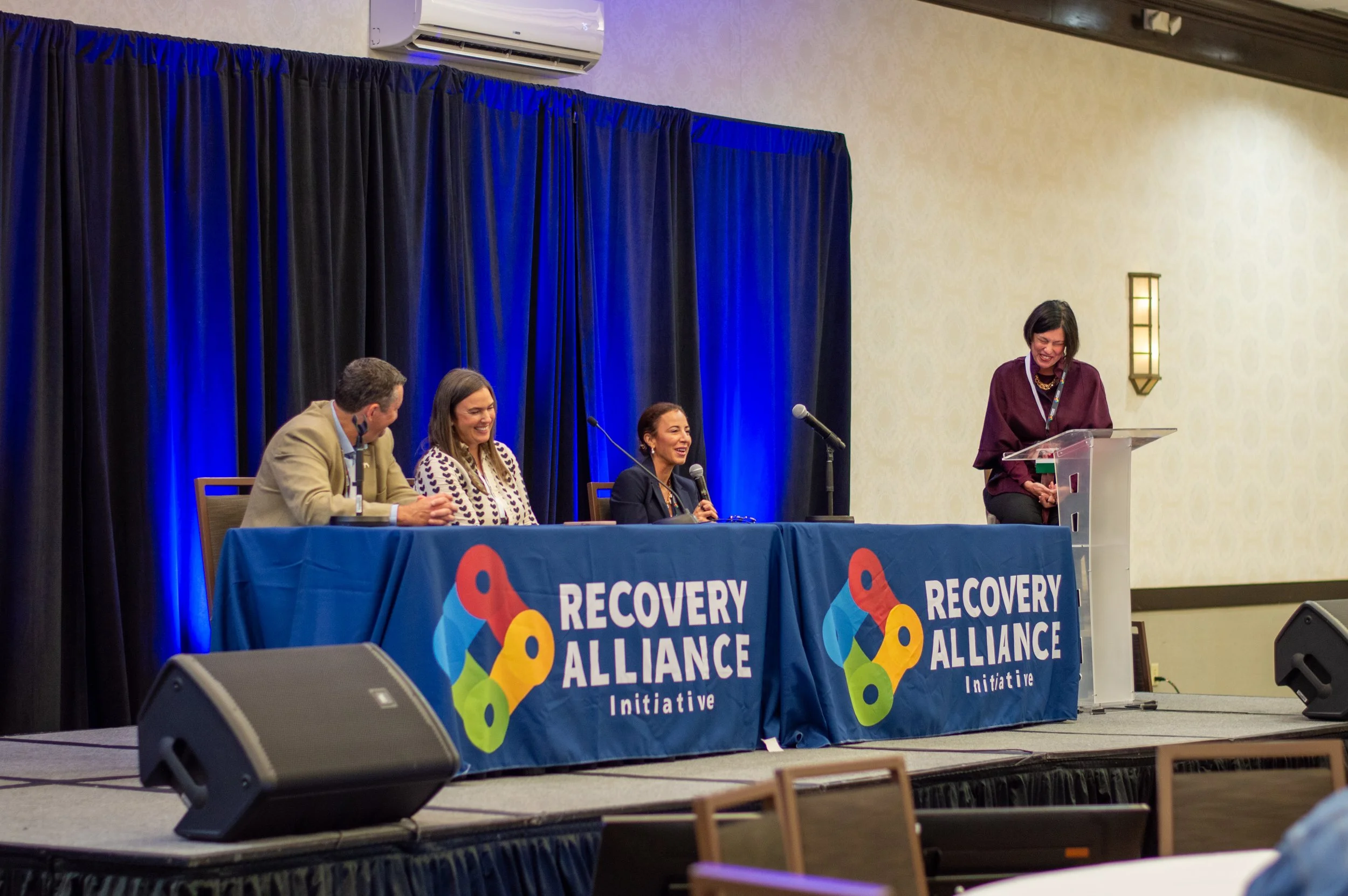 Four people seated behind a table with a blue tablecloth that reads 'Recovery Alliance Initiative,' on a stage with a woman standing at a podium speaking into a microphone, black curtains in the background, beige walls, and a speaker on the floor.