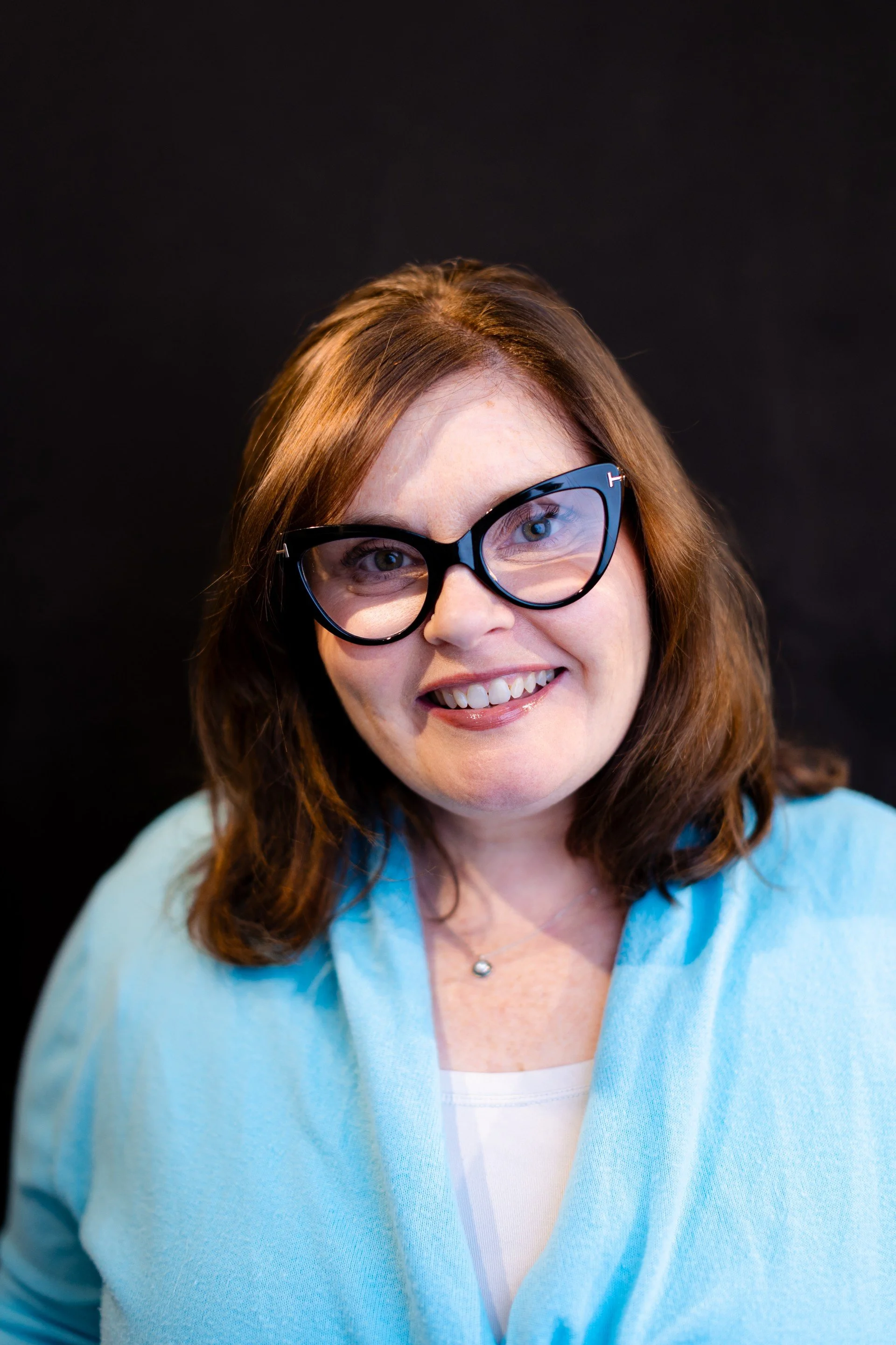 A woman with shoulder-length brown hair, wearing black eyeglasses, a light blue cardigan, and a white top, smiling against a black background.