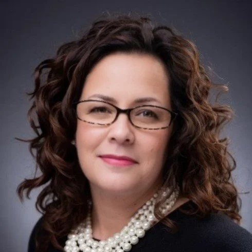A woman with curly brown hair, wearing glasses, a black top, and a pearl necklace, looking at the camera with a slight smile against a dark background.