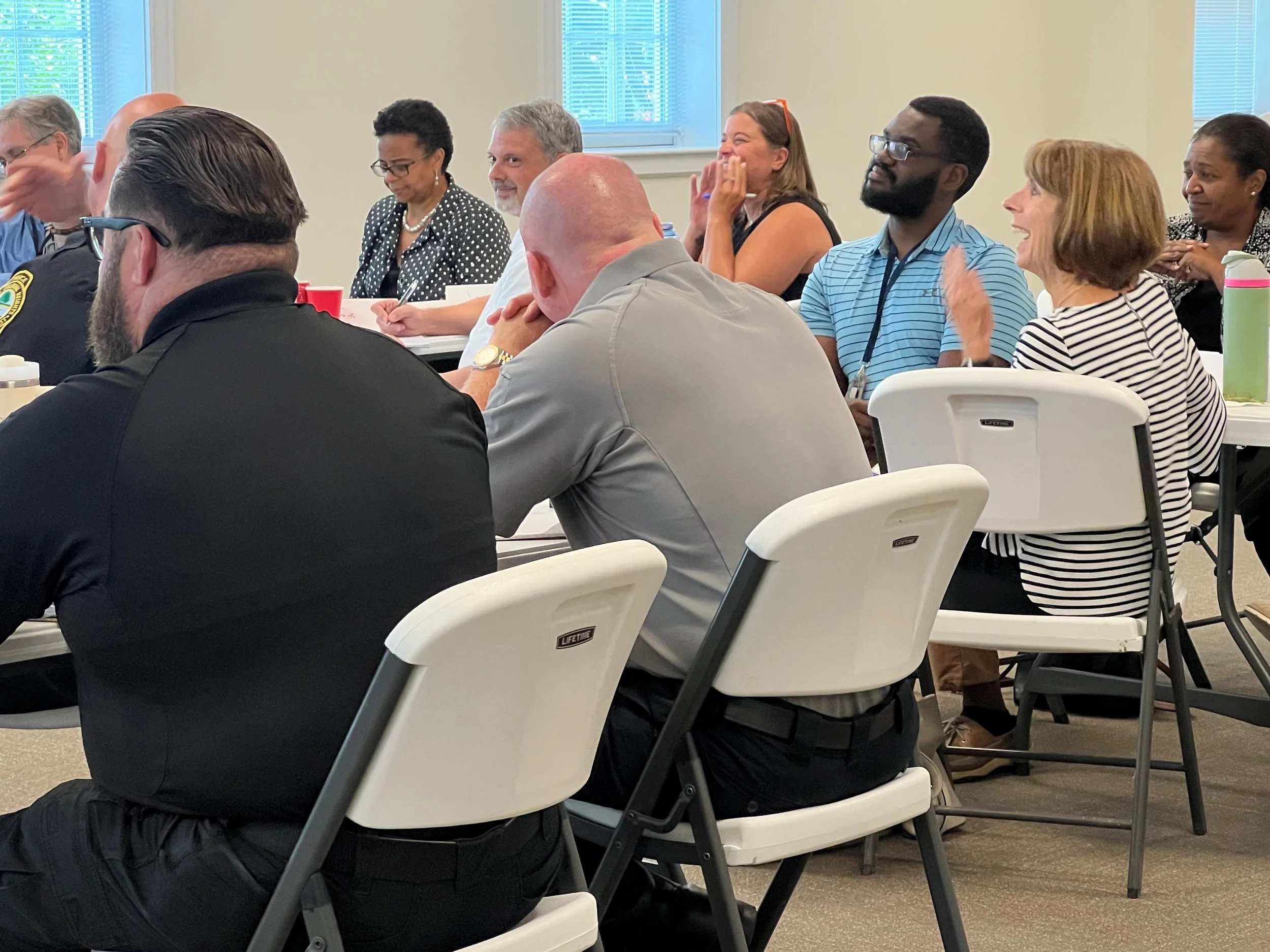A group of people sitting at tables, engaging and listening in a meeting or conference room with blinds and windows in the background.