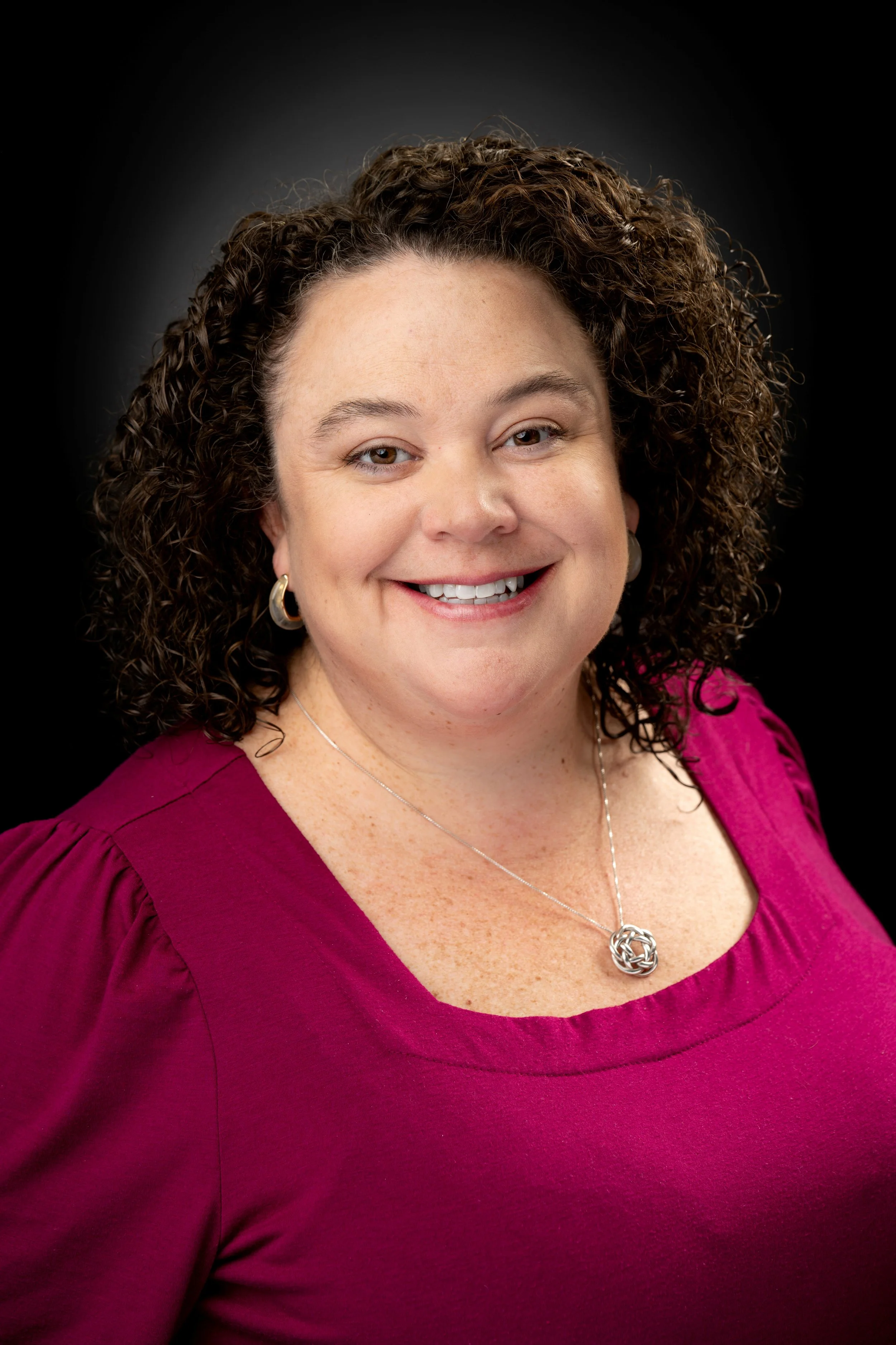 A woman with curly brown hair, wearing a magenta top, a silver necklace, and hoop earrings, smiling against a black background.