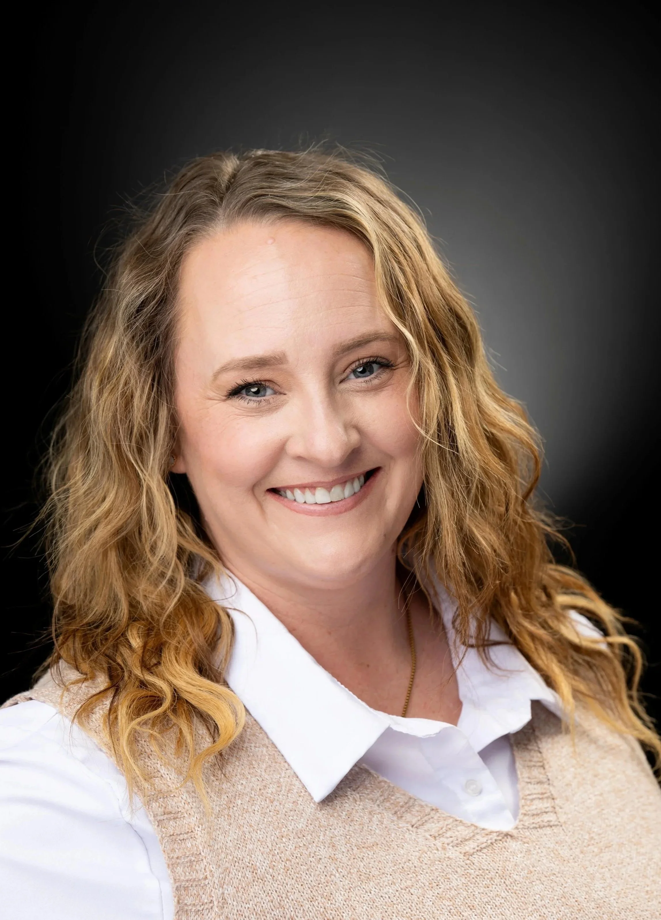 A smiling woman with long, wavy, blonde hair wearing a white shirt and beige vest against a dark background.