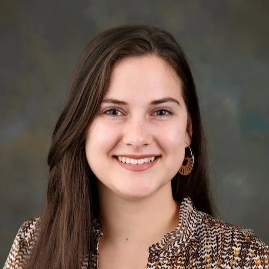 A young woman with long brown hair, smiling, wearing earrings and a patterned top against a plain background.