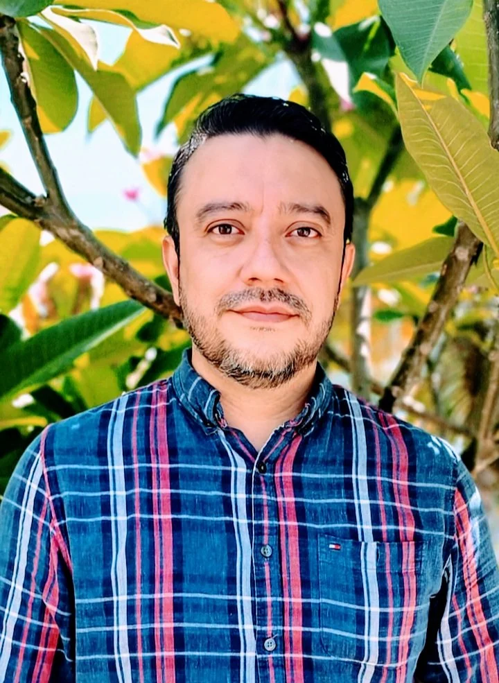 A man with dark hair and a beard smiling outdoors in front of green and yellow leaves, wearing a blue plaid shirt.