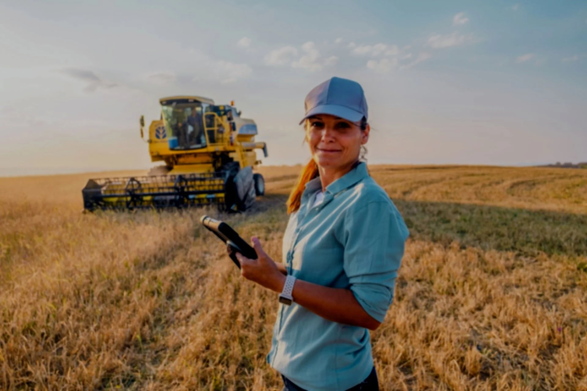 A woman standing in a field holding a tablet, with a large yellow harvest machine working in the background during sunset.
