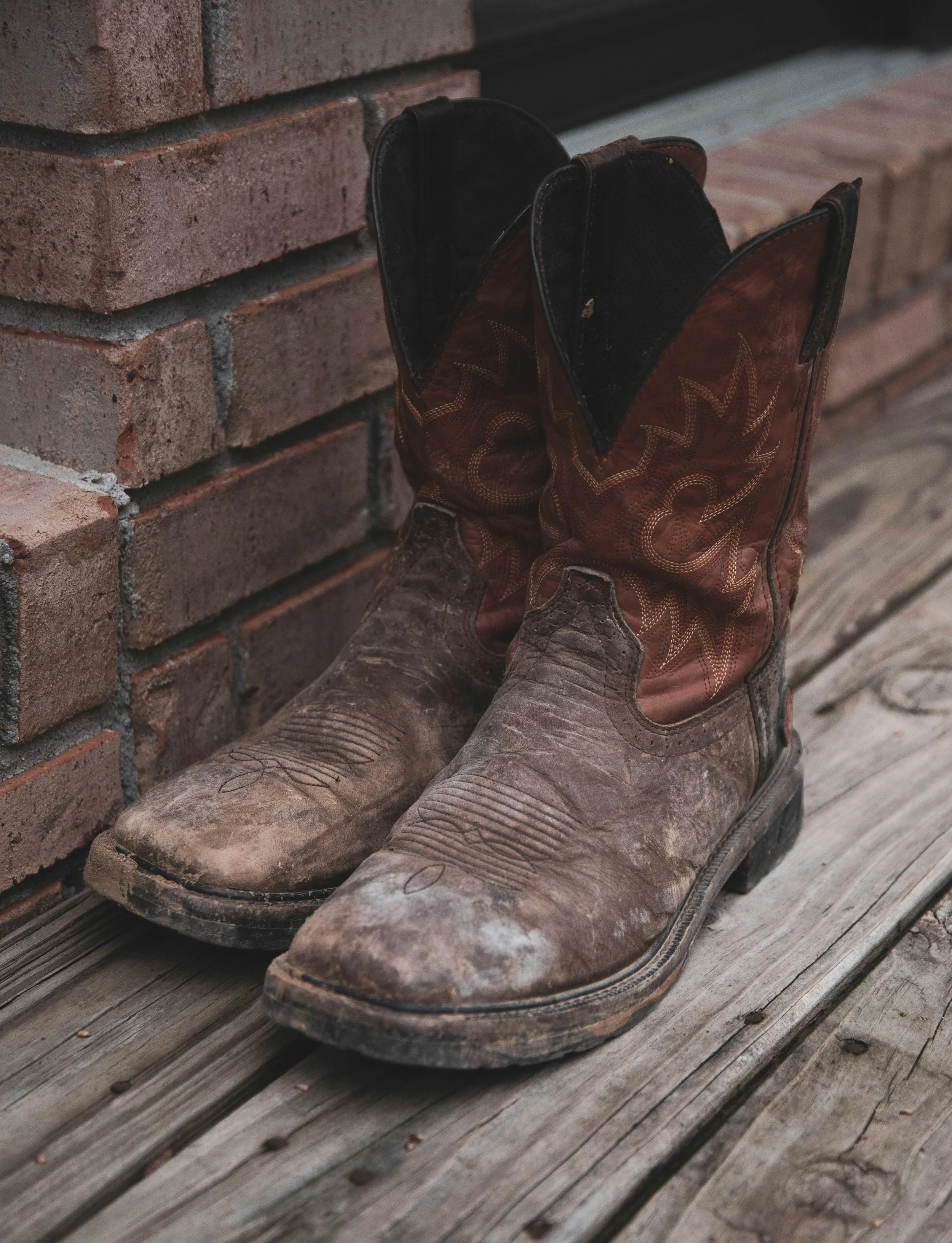 A pair of old, muddy cowboy boots resting against a brick wall on a wooden deck.