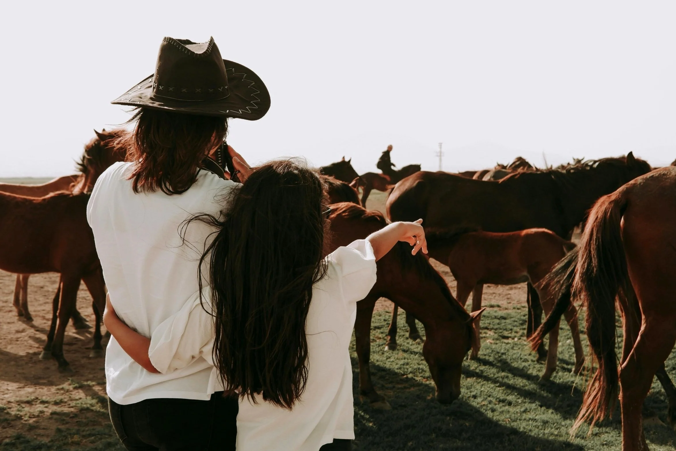 Two people, one adult and one child, are standing among grazing horses on a farm or ranch. The adult is wearing a cowboy hat. The child has long dark hair and is pointing at a horse. There are multiple horses in the background, with some grazing on grass and others standing on the dirt ground.