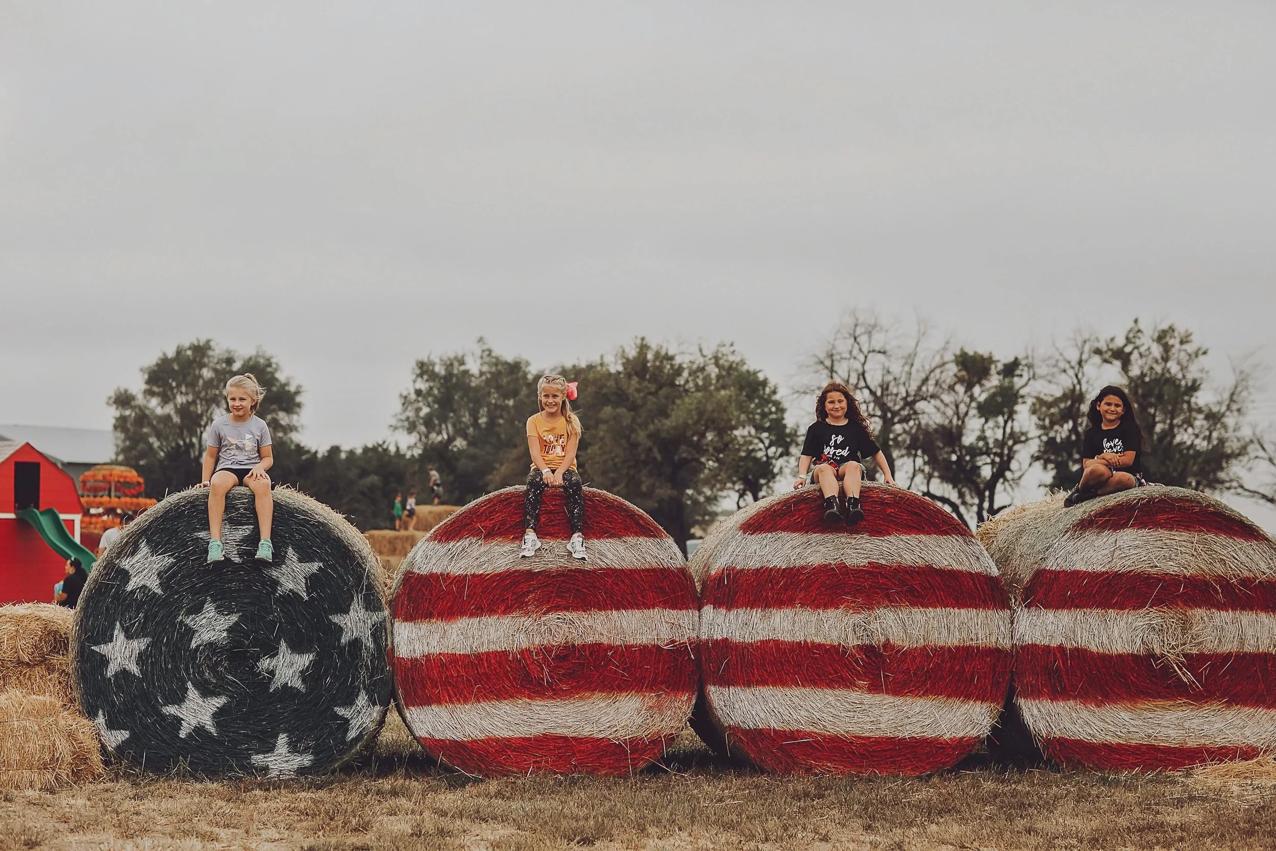 Four children sitting on large hay bales decorated with American flag designs at a rural farm.