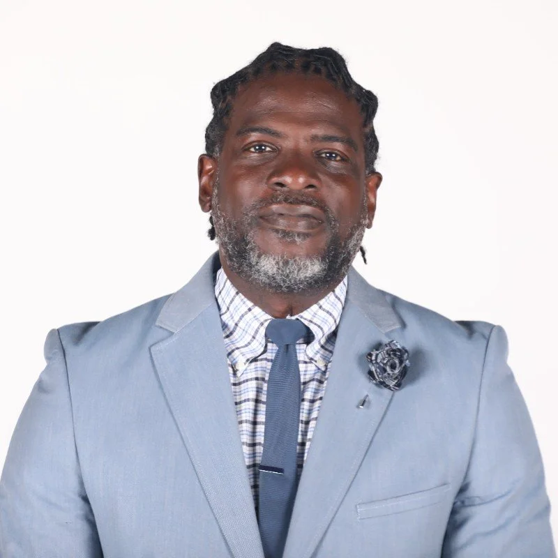 A well-dressed man with dreadlocks and a beard wearing a light blue suit, checkered shirt, and dark tie, posing against a plain white background.