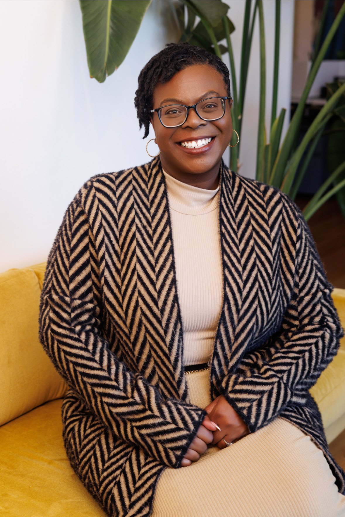 A smiling woman with glasses and hoop earrings sitting on a yellow couch indoors, wearing a cream turtleneck and a patterned blazer, with large green houseplants in the background.