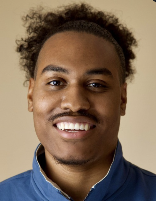 Portrait of a young man with curly hair, wearing a blue jacket, smiling against a plain beige background.