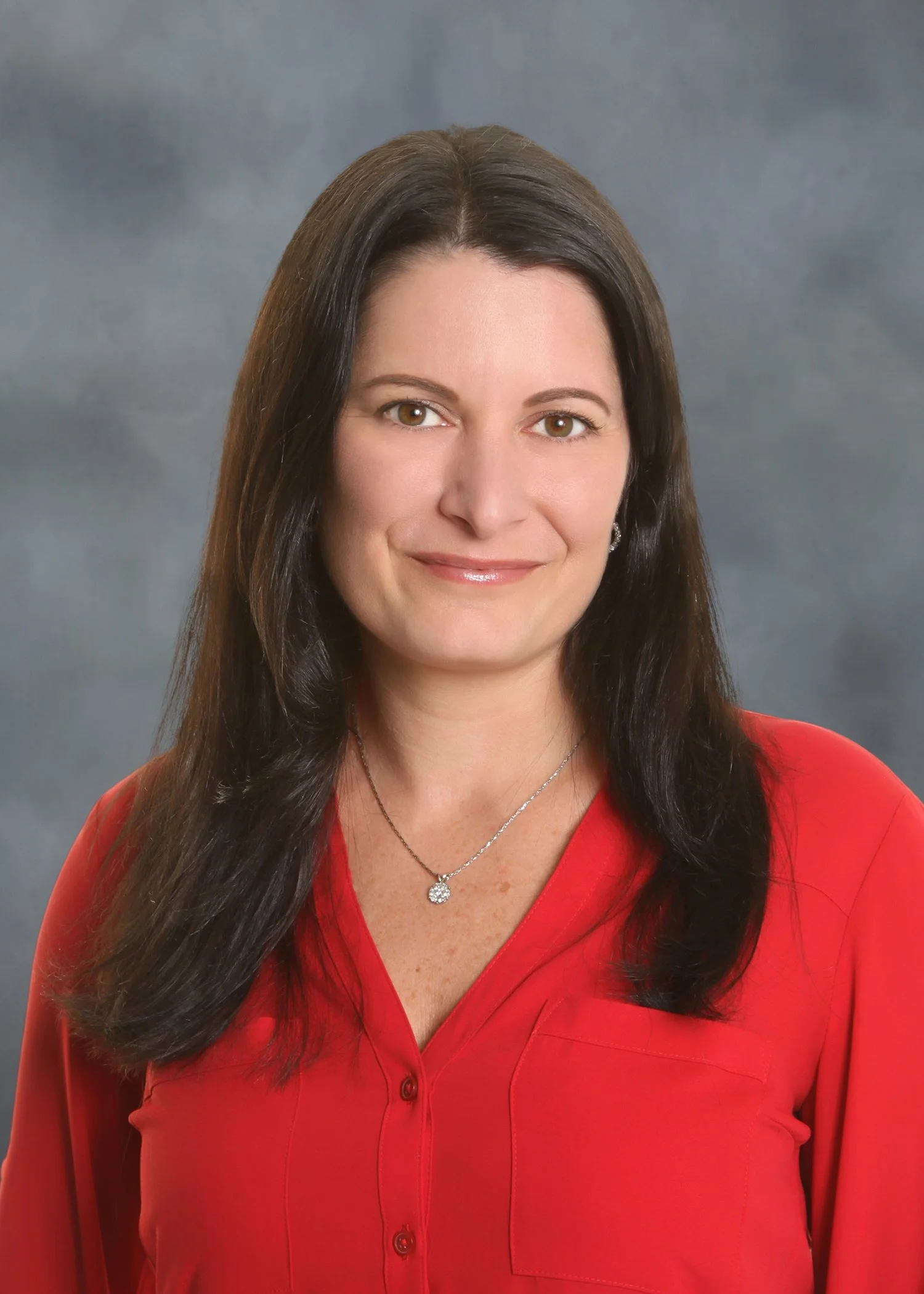Headshot of a woman with long dark brown hair wearing a red blouse and a silver necklace, against a gray background.