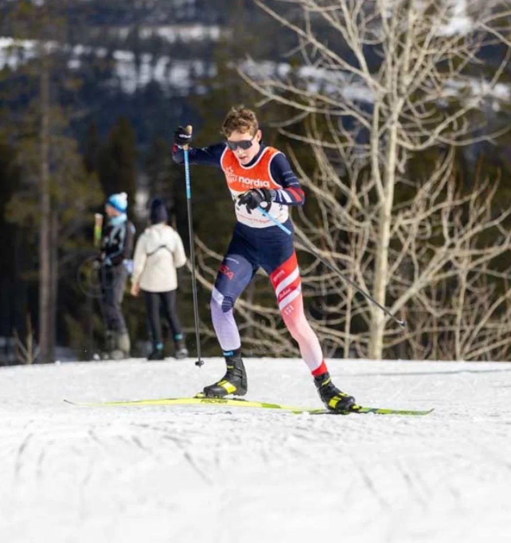Un jeune athlète en ski de fond en compétition sur piste enneigée, portant une combinaison de sport colorée, avec d'autres skieurs en arrière-plan et des arbres sans feuilles.