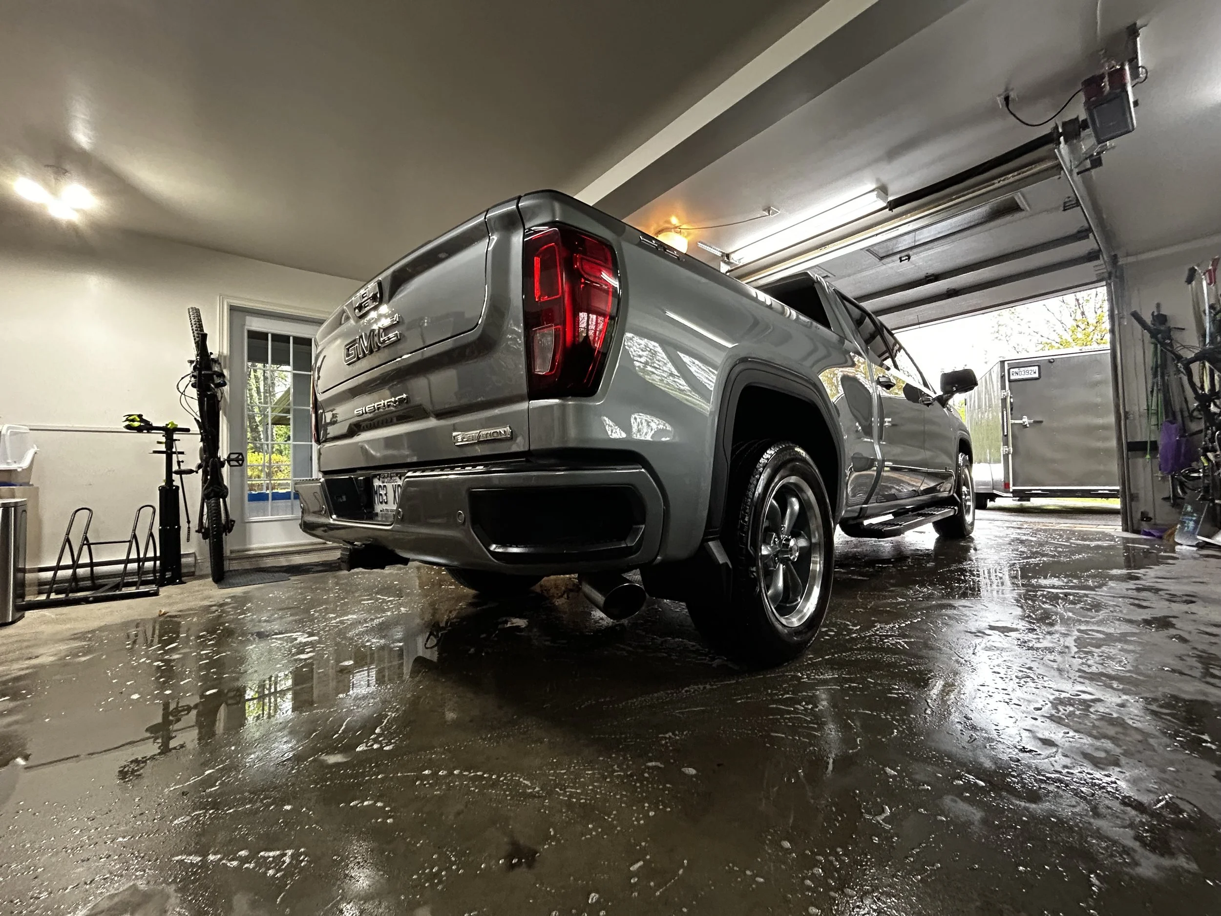 Véhicule GMC Sierra dans un garage, vue de l'arrière et côté, le sol mouillé reflète la voiture, porte de garage ouverte sur l'extérieur.