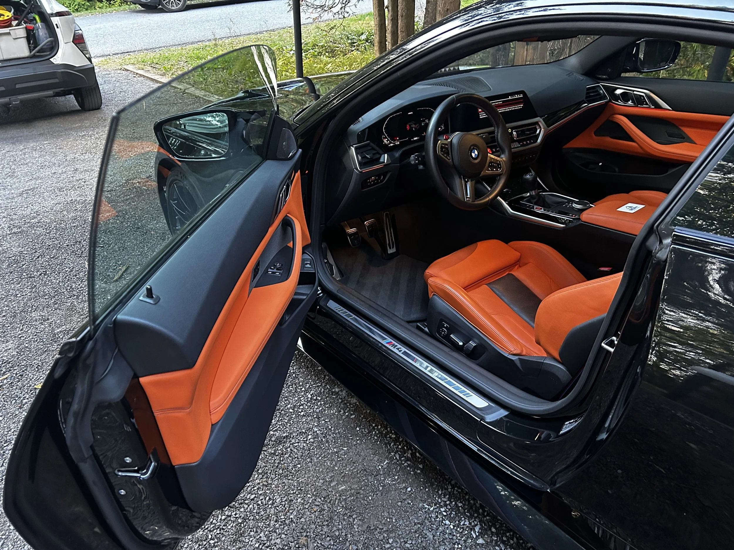 Intérieur d'une voiture noire BMW M2 avec des sièges en cuir orange, vue de l'ouverture de la porte côté conducteur.