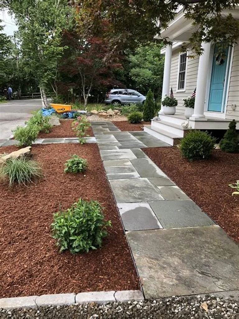 Stone walkway leading to the front porch of a house with white columns and blue front door, surrounded by landscaped garden beds with small bushes and mulch.