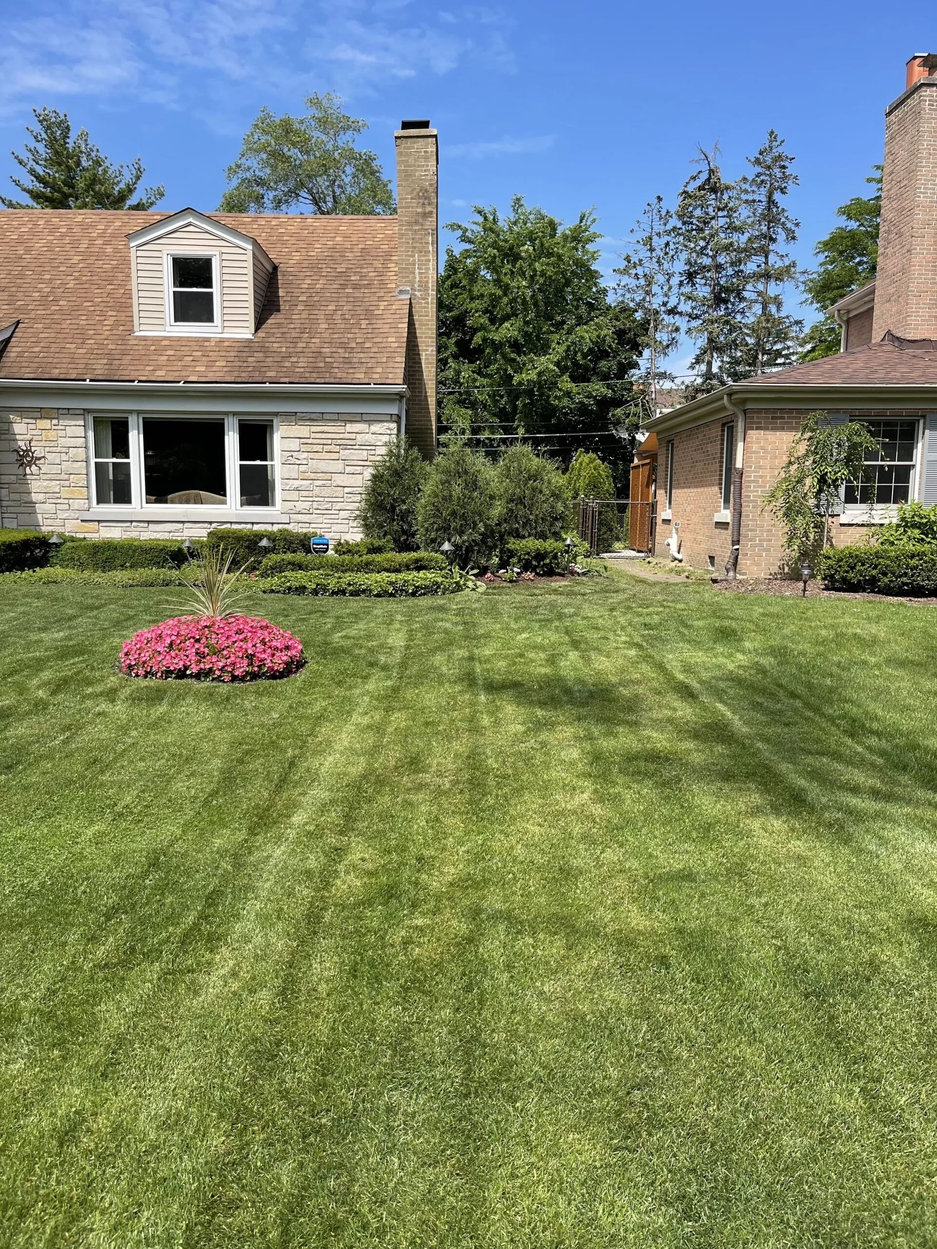 A well-maintained house with a stone and brick exterior, a large front window, a small dormer window, and a chimney. There is a lush green lawn with a circular flower bed containing pink flowers in the front yard. Trees and bushes surround the house, with a clear blue sky overhead.