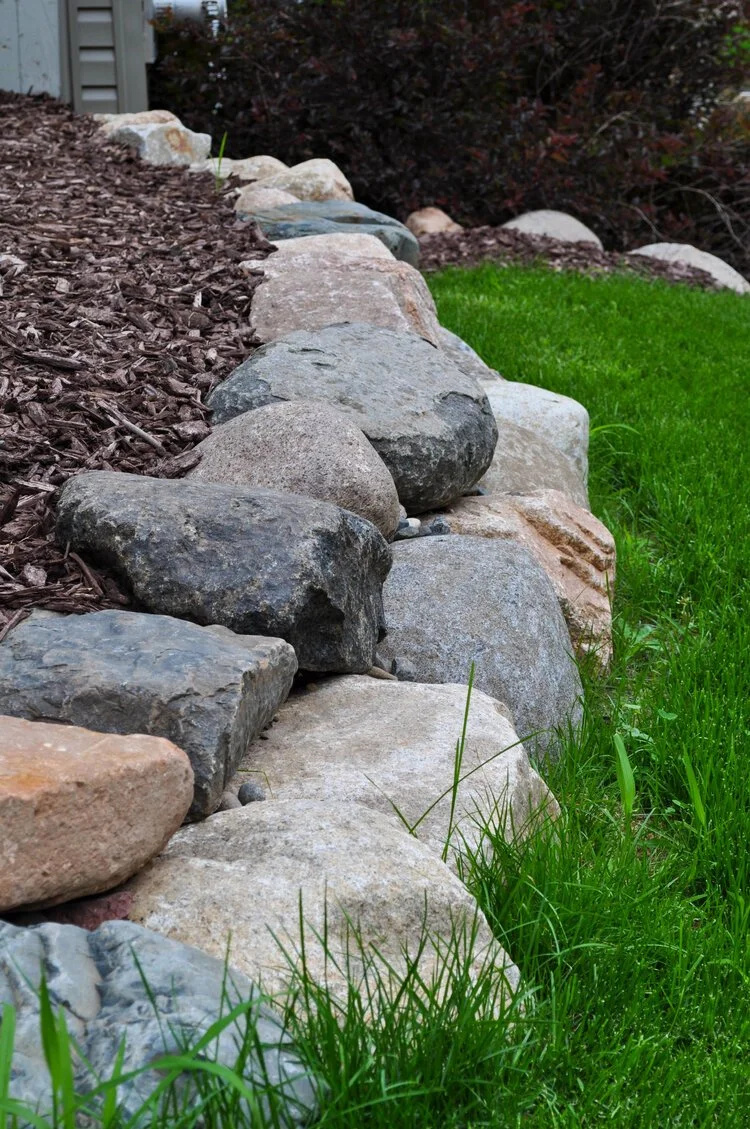 A garden landscape with a curved border of large gray and tan rocks separating a mulch-covered area from a lush green grass lawn, with dark shrubbery in the background.