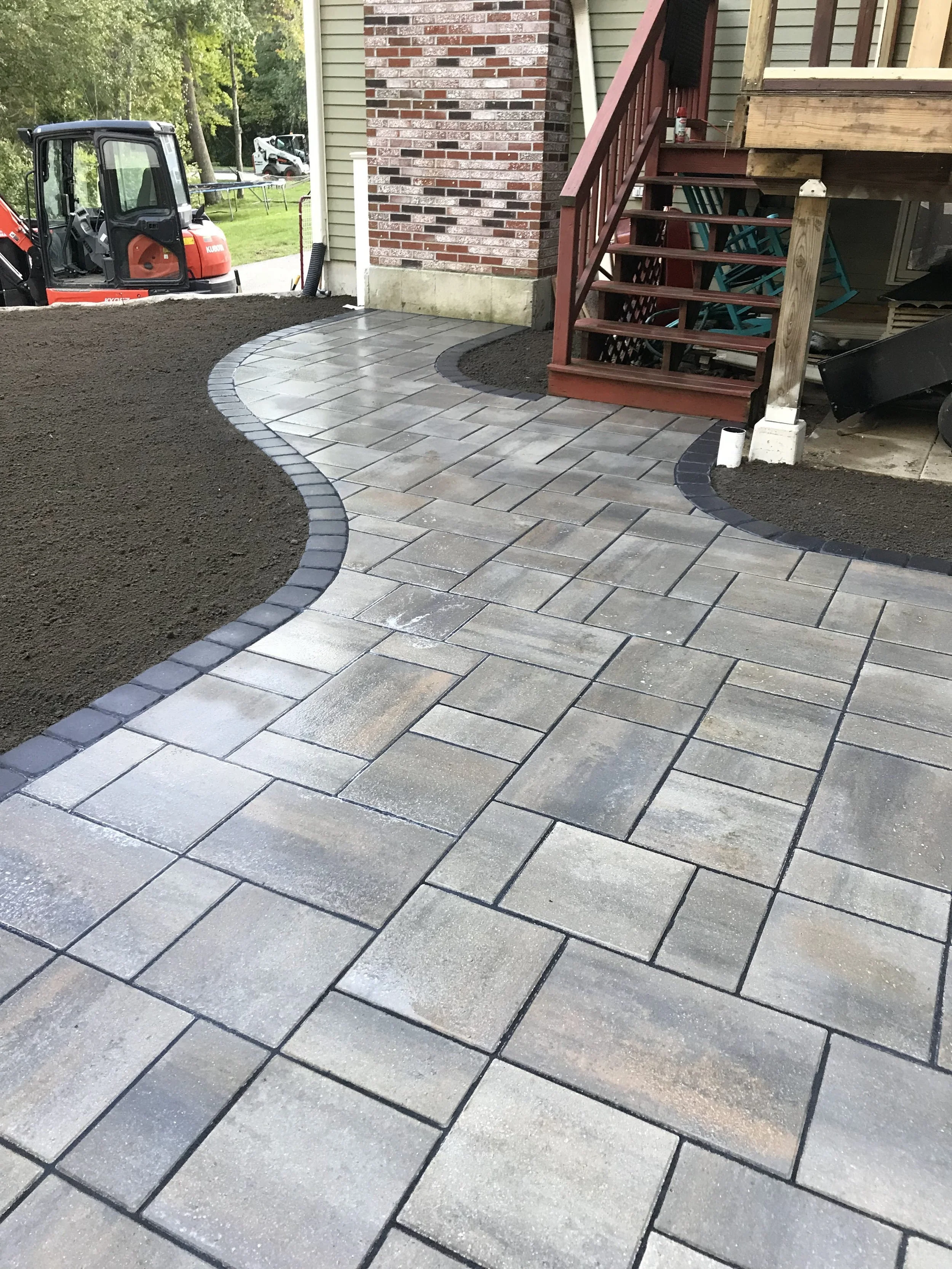 Newly installed stone pathway winding around a house with dark borders, concrete steps with a reddish stair railing, and construction equipment nearby.