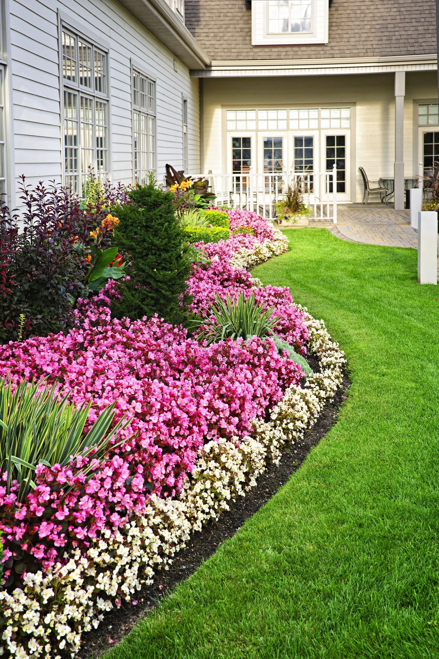 A well-maintained backyard garden with colorful pink, white, and purple flowers along a green lawn, house with white exterior, multiple windows, and a patio with outdoor furniture.