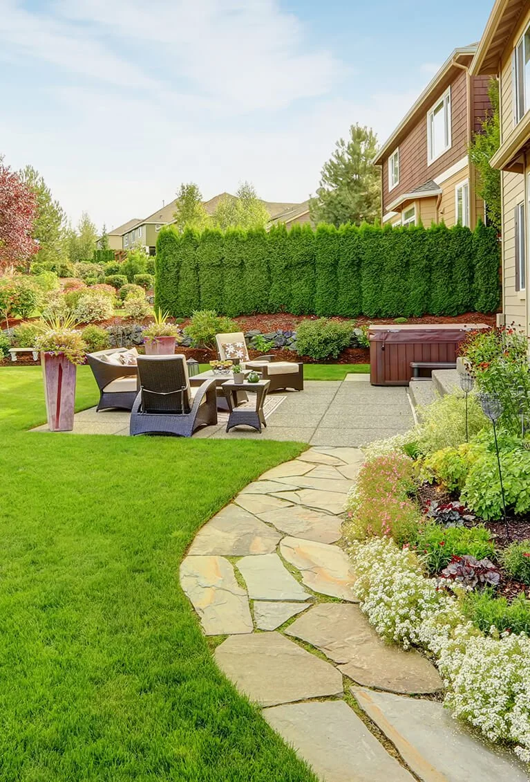 A well-maintained backyard featuring a stone pathway, green lawn, outdoor seating area with chairs and small tables, lush shrubs, colorful flowers, and a tall hedge providing privacy, with houses visible in the background.