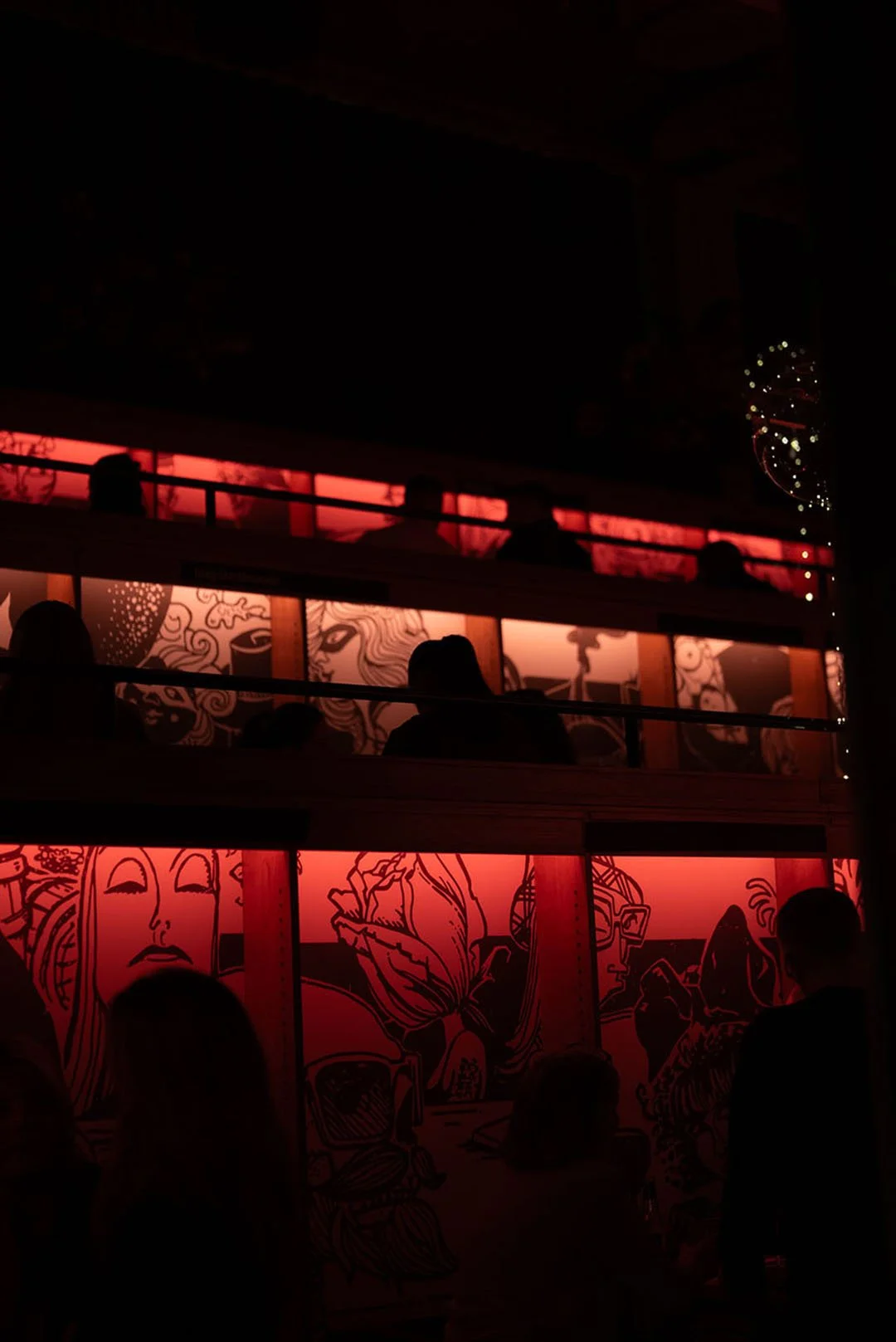 Mänskor på en balkong mittemot en vägg med tecknade konstverk, belysning i rött och rosa nyanser.
