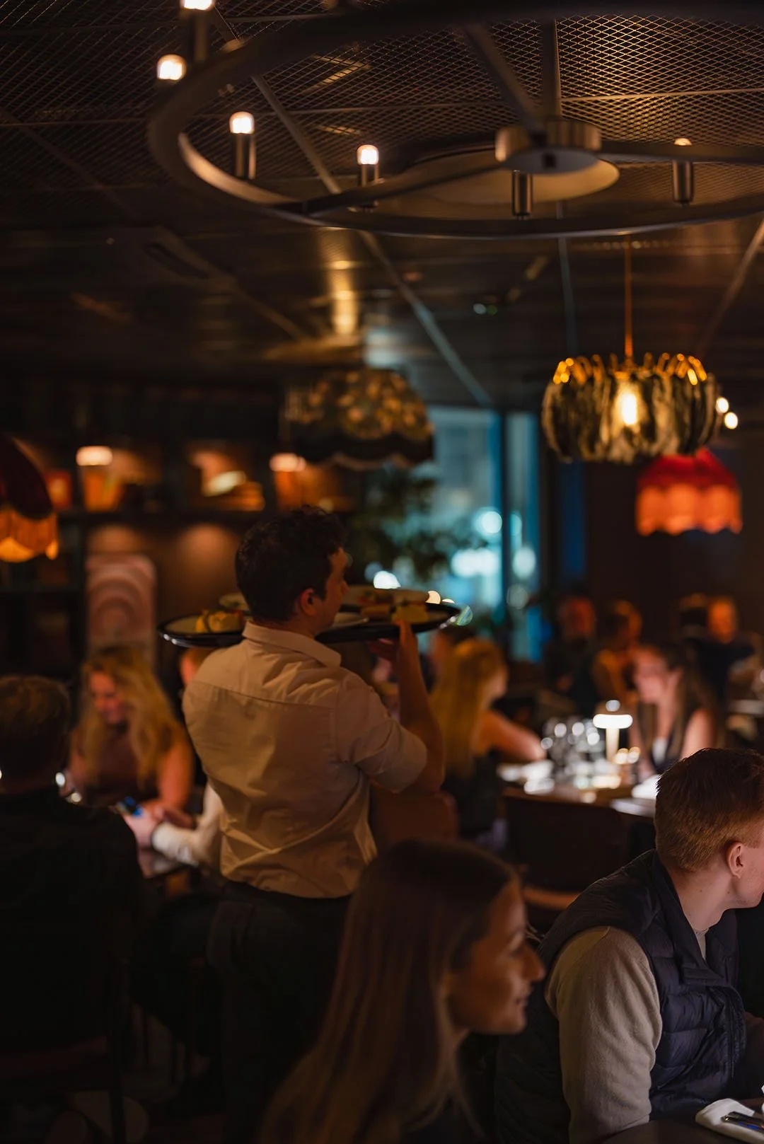En livlig restaurang med många gäster som äter och samtalar, medan en servitör bär en bricka med mat.