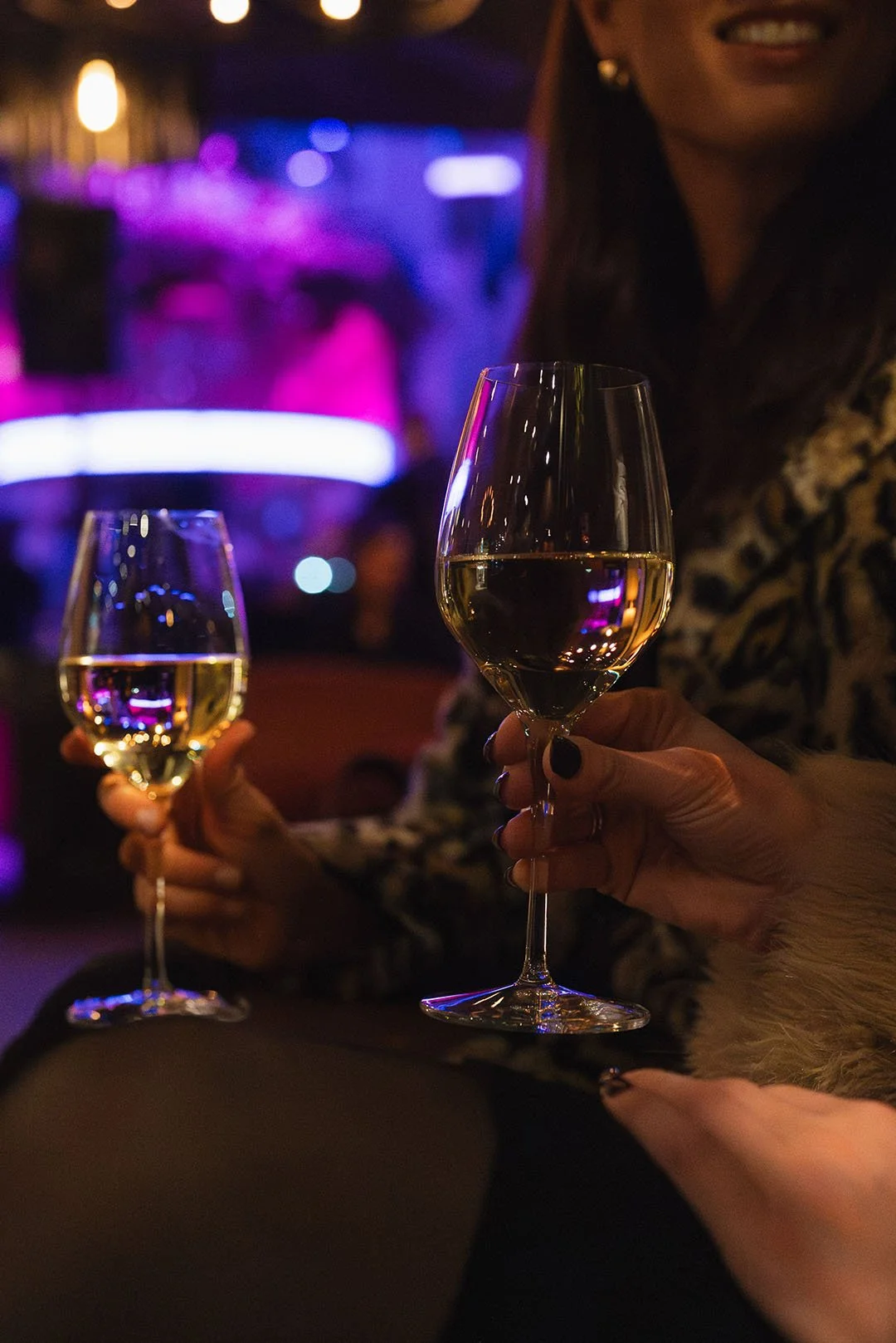 Två personer som håller i vinglas fyllda med vitt vin i en bar eller klubbmiljö med färgglada ljus i bakgrunden.