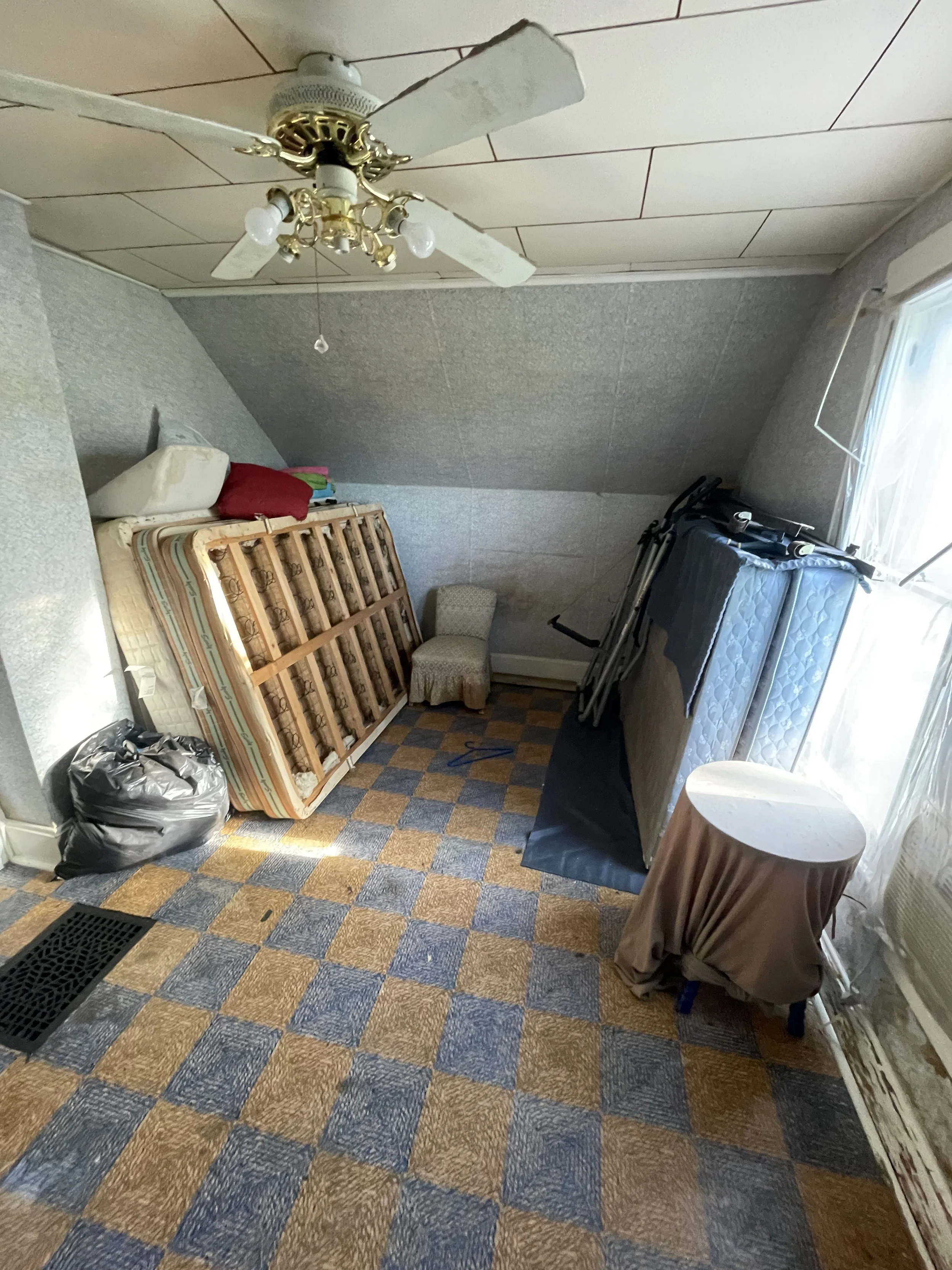 A cluttered room with a sloped ceiling, a ceiling fan, a mattress leaning against the wall, a small table with a cloth cover, a chair, and folded bed frames or furniture. The room has a checkered carpet and a window with curtains.