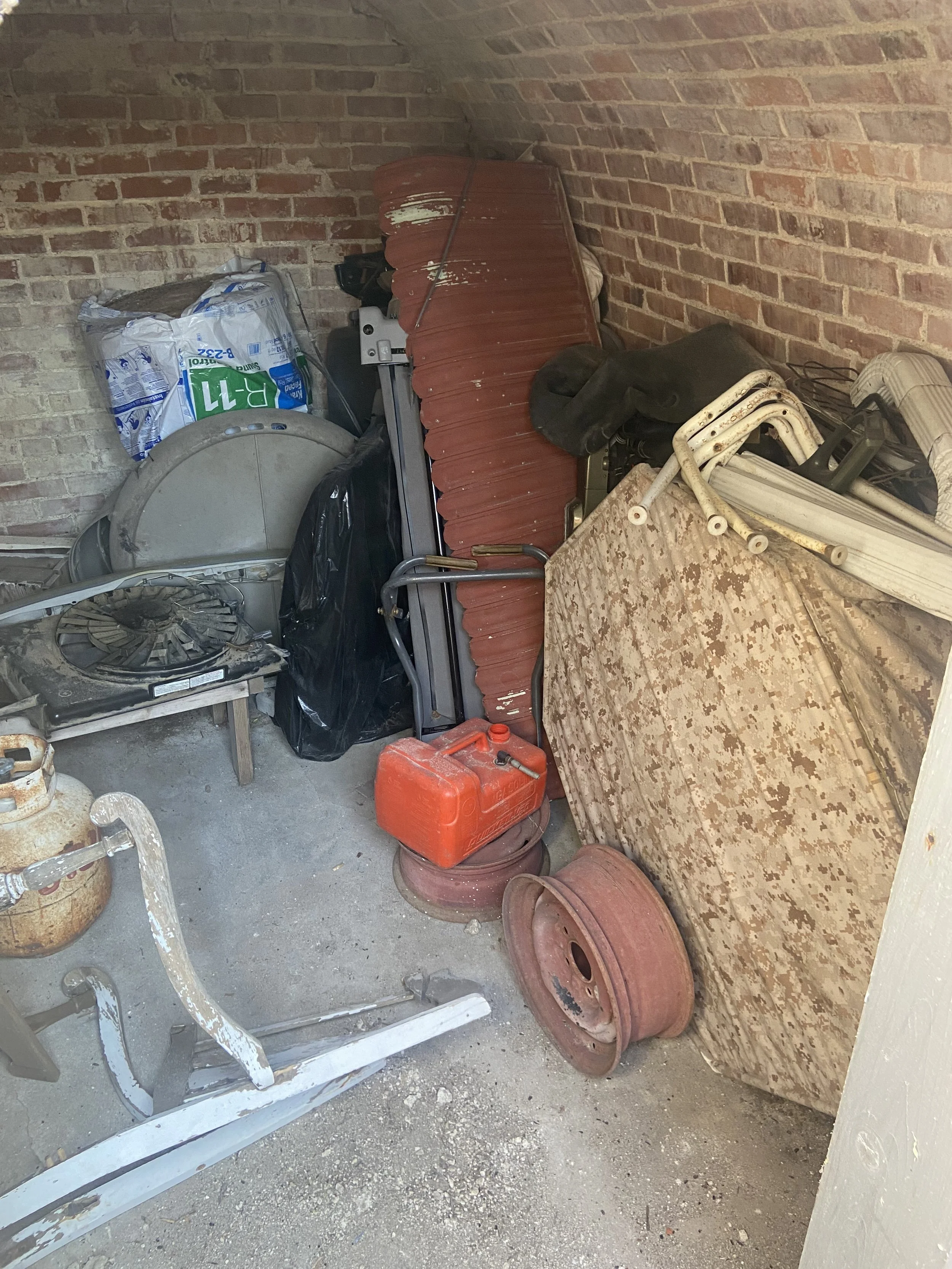 Cluttered garage corner with rusted tires, old patio furniture, plastic containers, tools, and miscellaneous items stacked against brick walls.