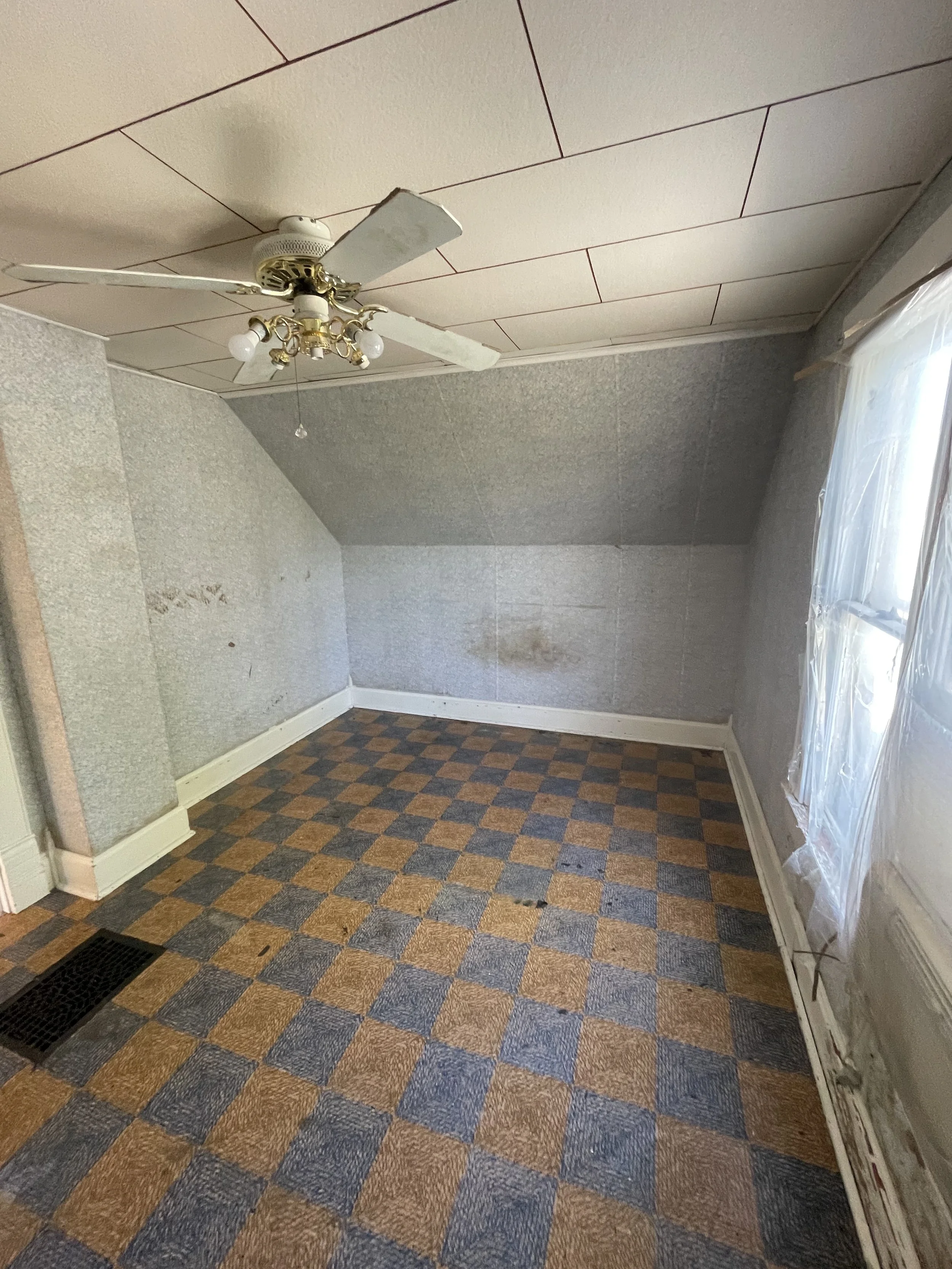Empty room with worn checkered carpet, textured gray walls, a white ceiling with a brass chandelier and ceiling fan, and a window covered with clear plastic sheeting.