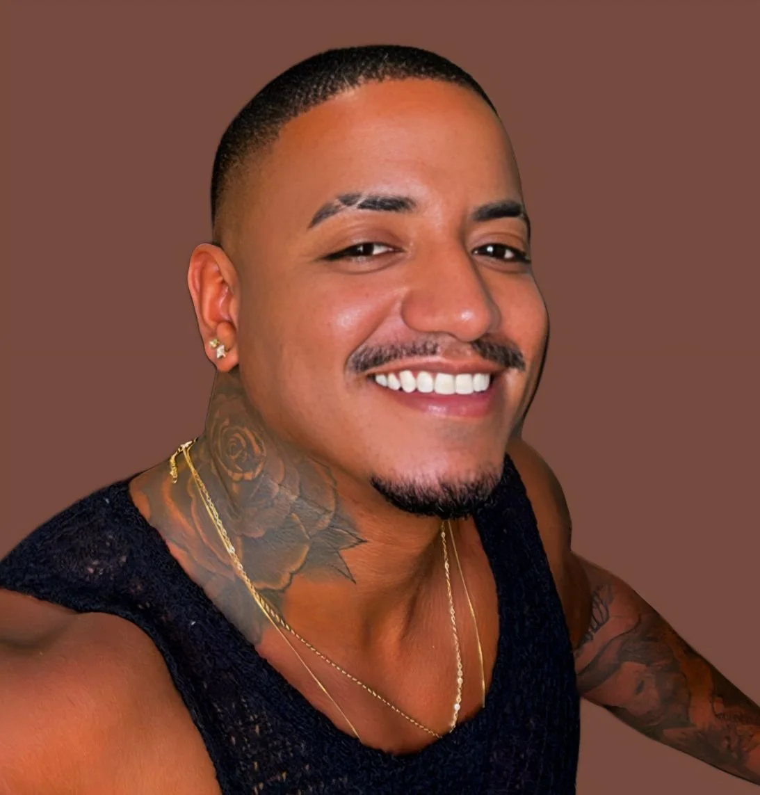 Homem sorrindo com cabelos curtos, tatuagens no pescoço e braço, usando uma blusa preta e acessórios dourados, contra fundo marrom.