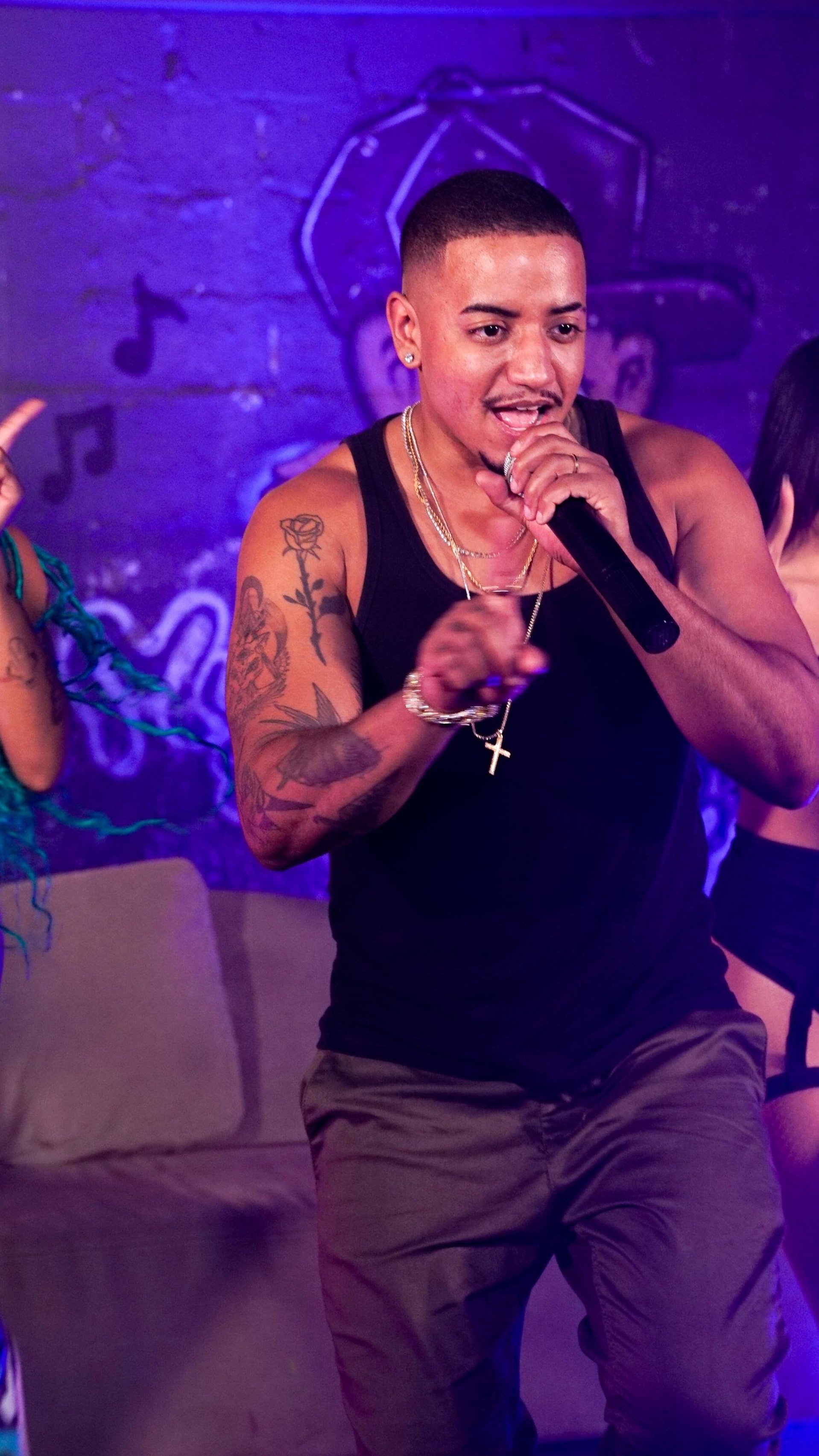 Homem com tatuagens usando camiseta preta, segurando um microfone, em uma cena de festa ou show com fundo roxo e pessoas ao redor.