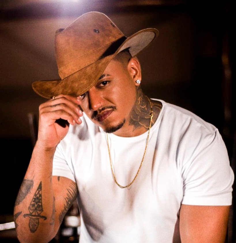 Homem com tatuagens, usando camiseta branca, com brincos, usando chapéu de cowboy marrom, posando com expressão confiante.