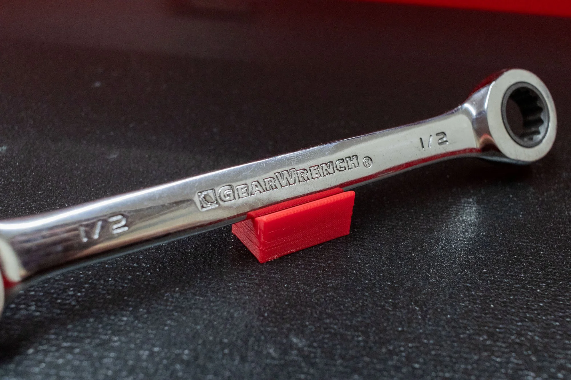 A metal gear wrench with size markings 1/2 inches, placed on a textured black surface with a red 3D-printed stand.