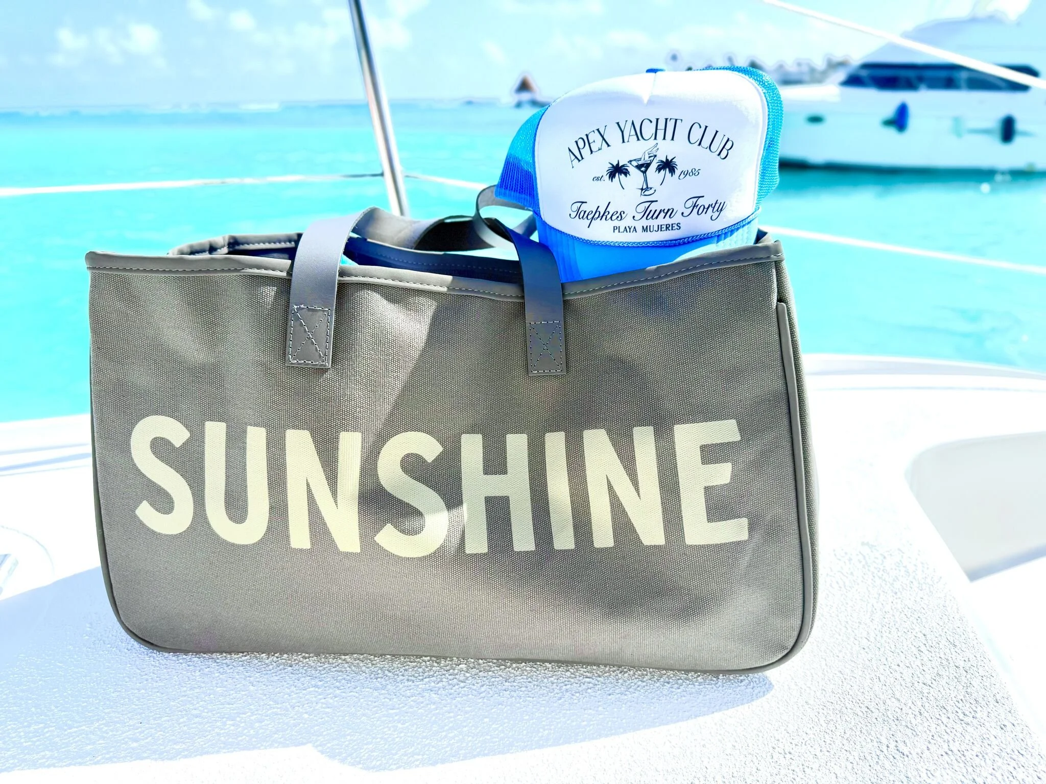 Gray bag with the word 'SUNSHINE' in large white letters on a boat deck with a turquoise sea and another boat in the background, also a blue and white cap behind the bag with 'APEX YACHT CLUB' text.