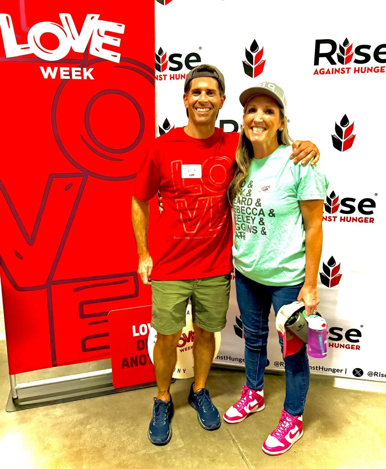 A man and a woman smiling and posing together at a Rise Against Hunger event, standing in front of a backdrop with the organization's logo. The man is wearing a red t-shirt and shorts, and the woman is in a mint green t-shirt, jeans, and pink sneaker