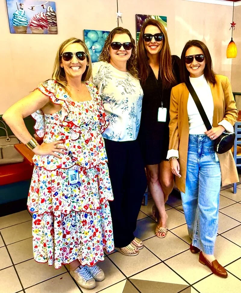 Four women wearing sunglasses and smiling, standing inside a restaurant or cafe with colorful artwork on the wall behind them.