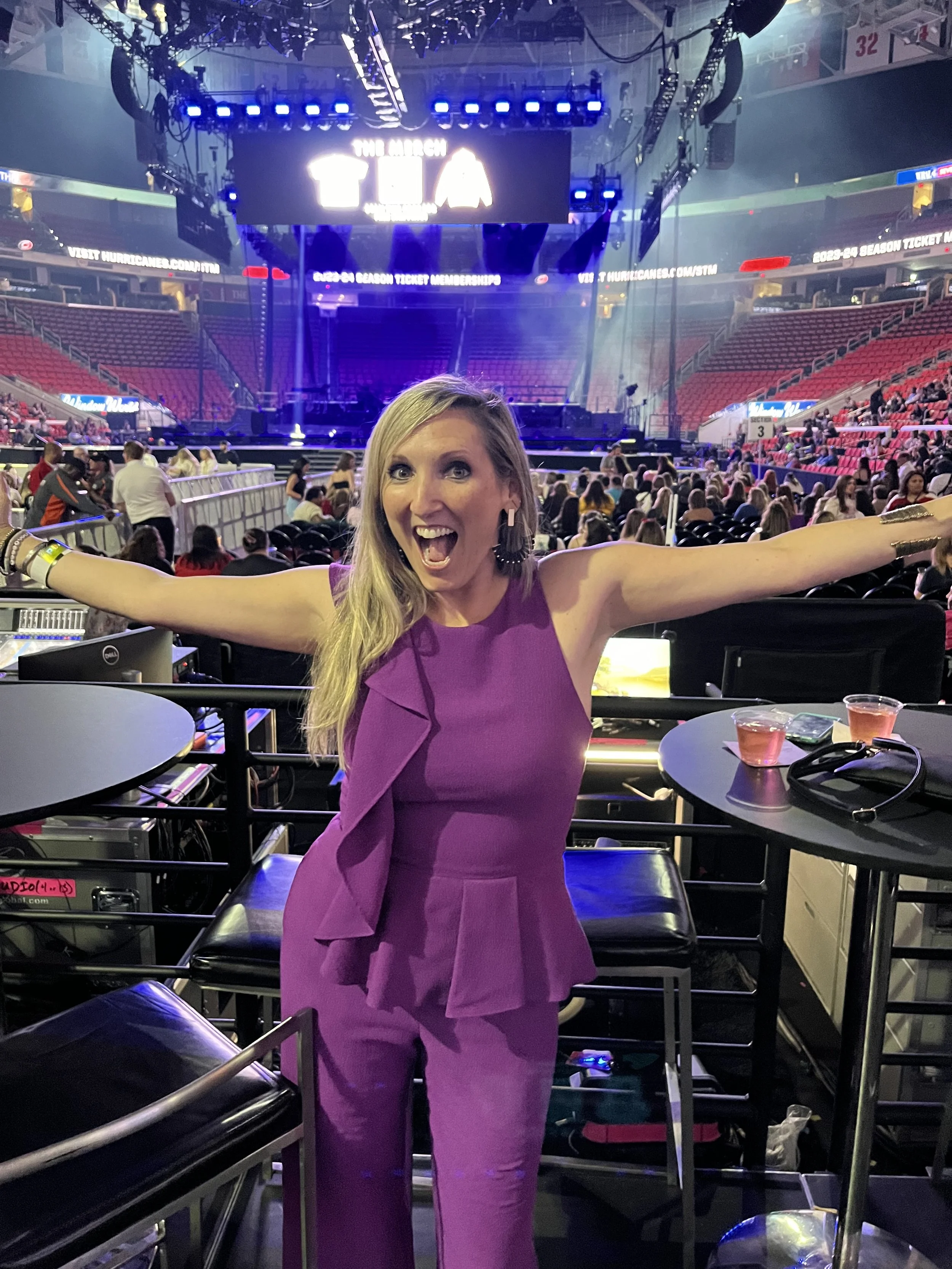 A woman in a purple outfit standing in front of a stage with her arms outstretched and smiling at a concert or event venue.