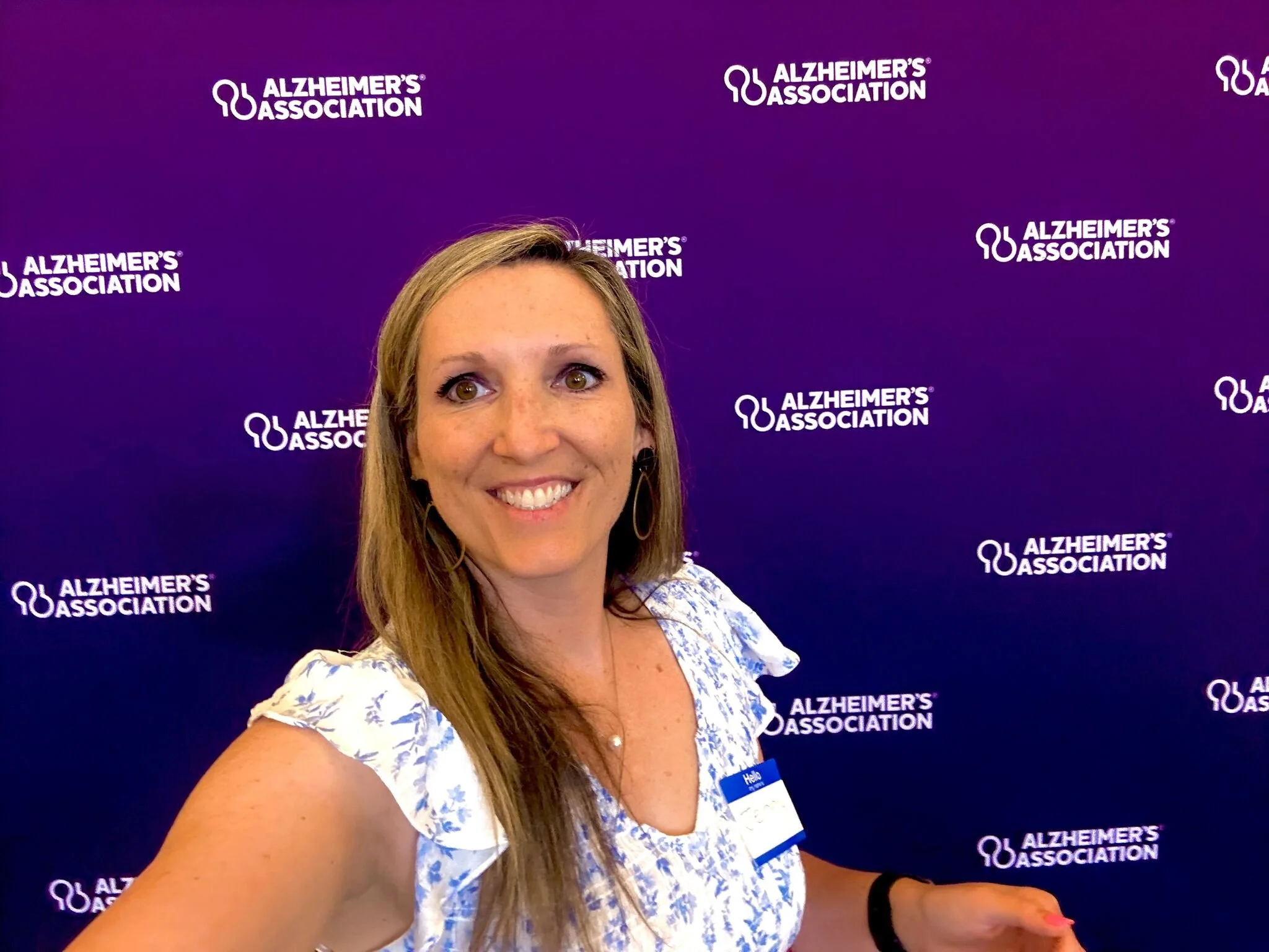 A woman with long blonde hair, wearing a white and blue patterned dress, smiling for a selfie in front of a purple Alzheimer's Association backdrop.