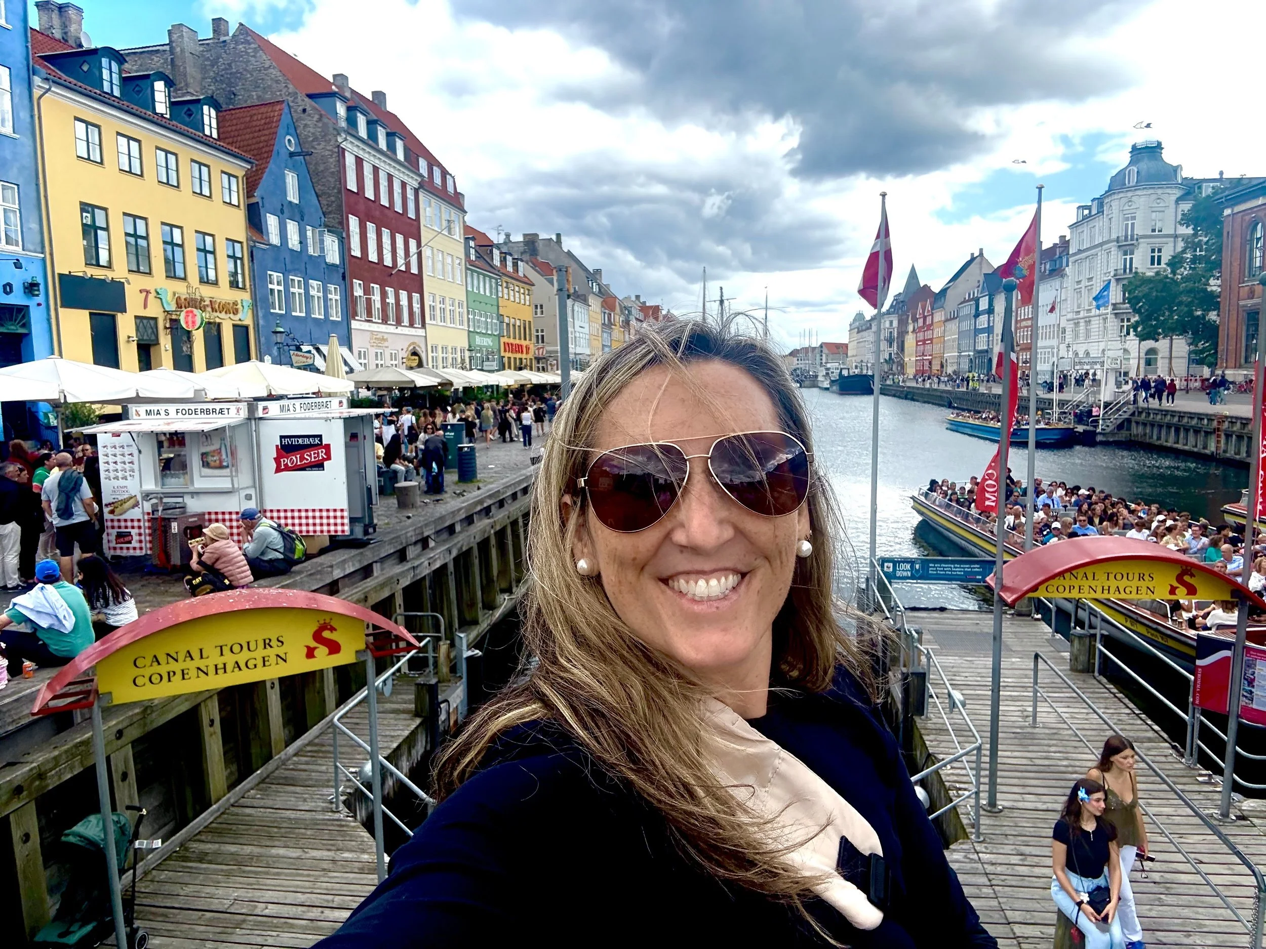 A woman taking a selfie at Nyhavn canal in Copenhagen, Denmark, with colorful historic buildings, boats, and tourists in the background.