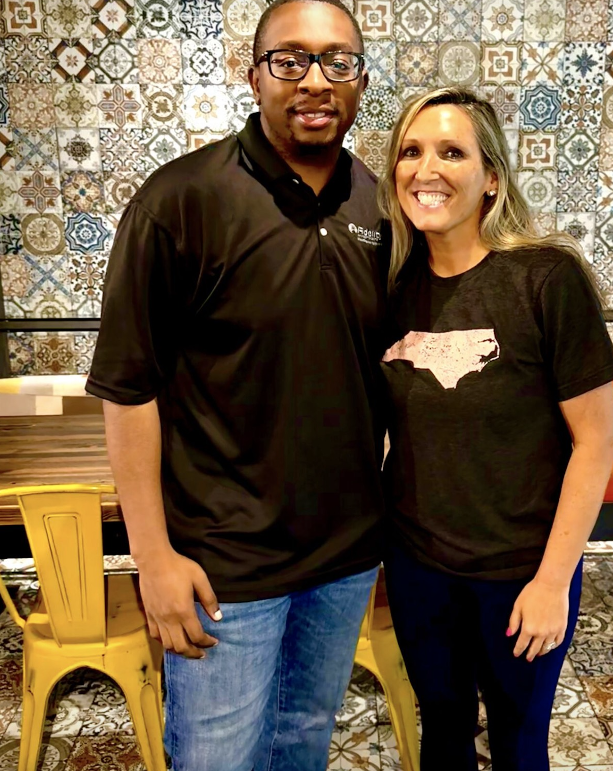 A man and a woman standing together indoors, smiling, with patterned tiles on the wall behind them.