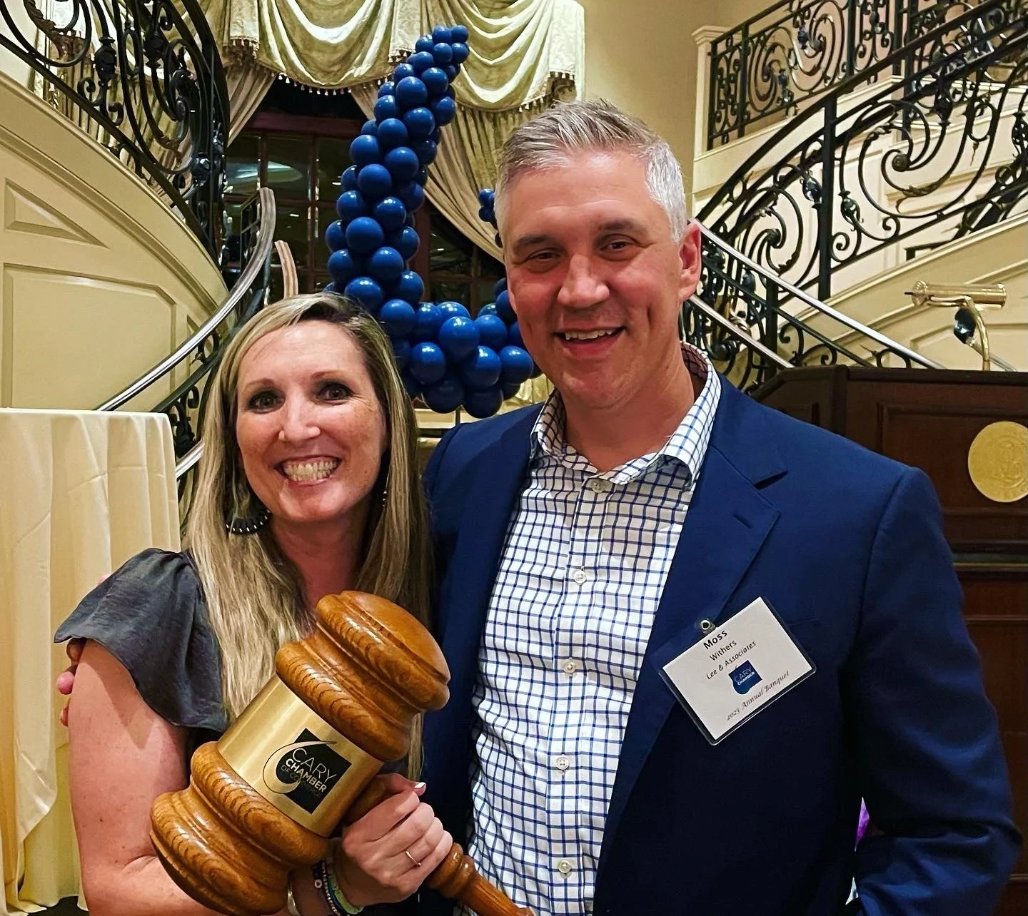 A woman and a man smiling for a photo at an event. The woman is holding a large gavel with a gold band that says 'CARY CHAMBER.' There are blue balloon decorations and a staircase with ornate railing in the background.