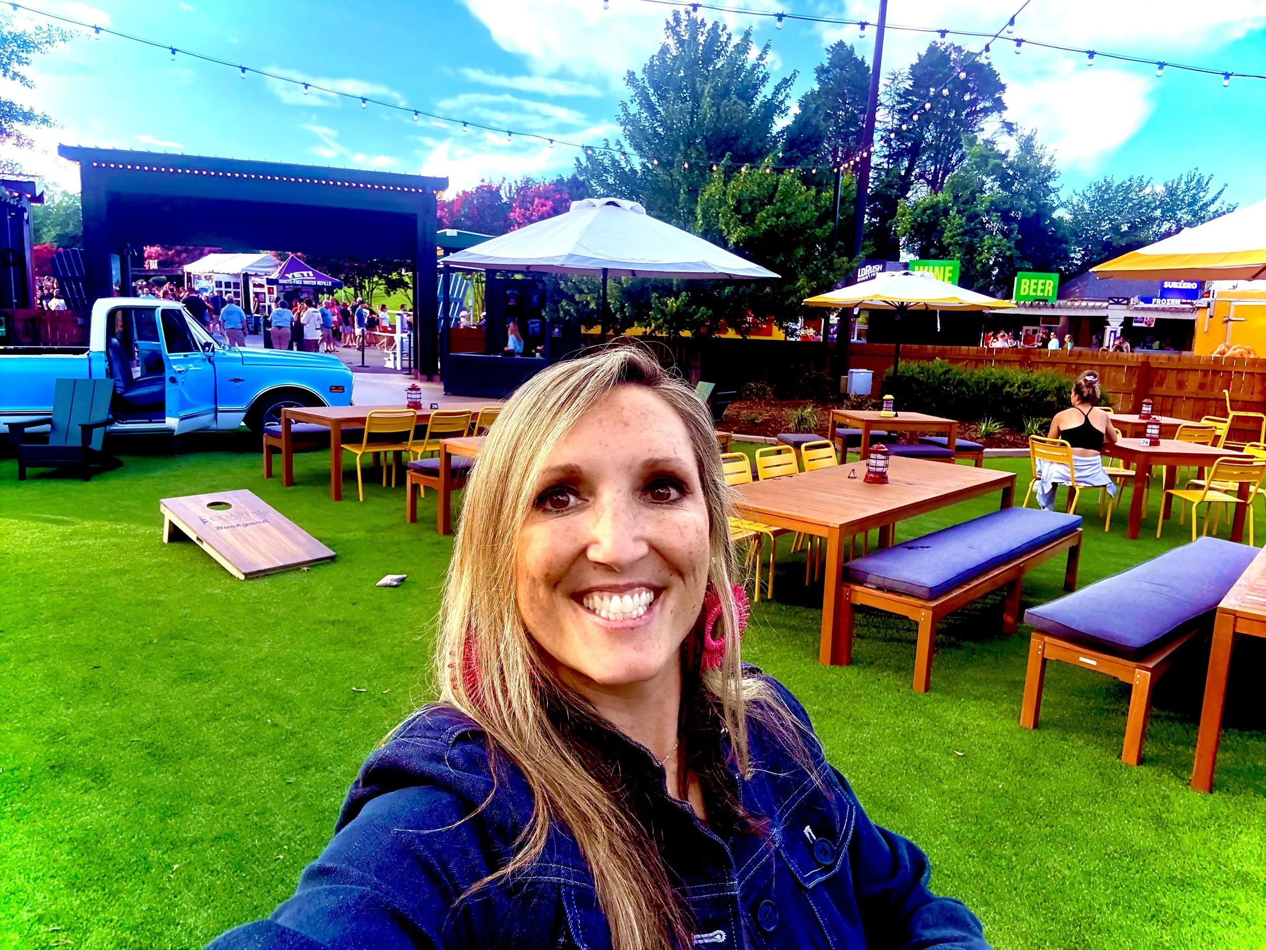 Jenny Hofler taking a selfie at an outdoor event with green grass, wooden tables and benches, umbrellas, and a stage in the background with a crowd.