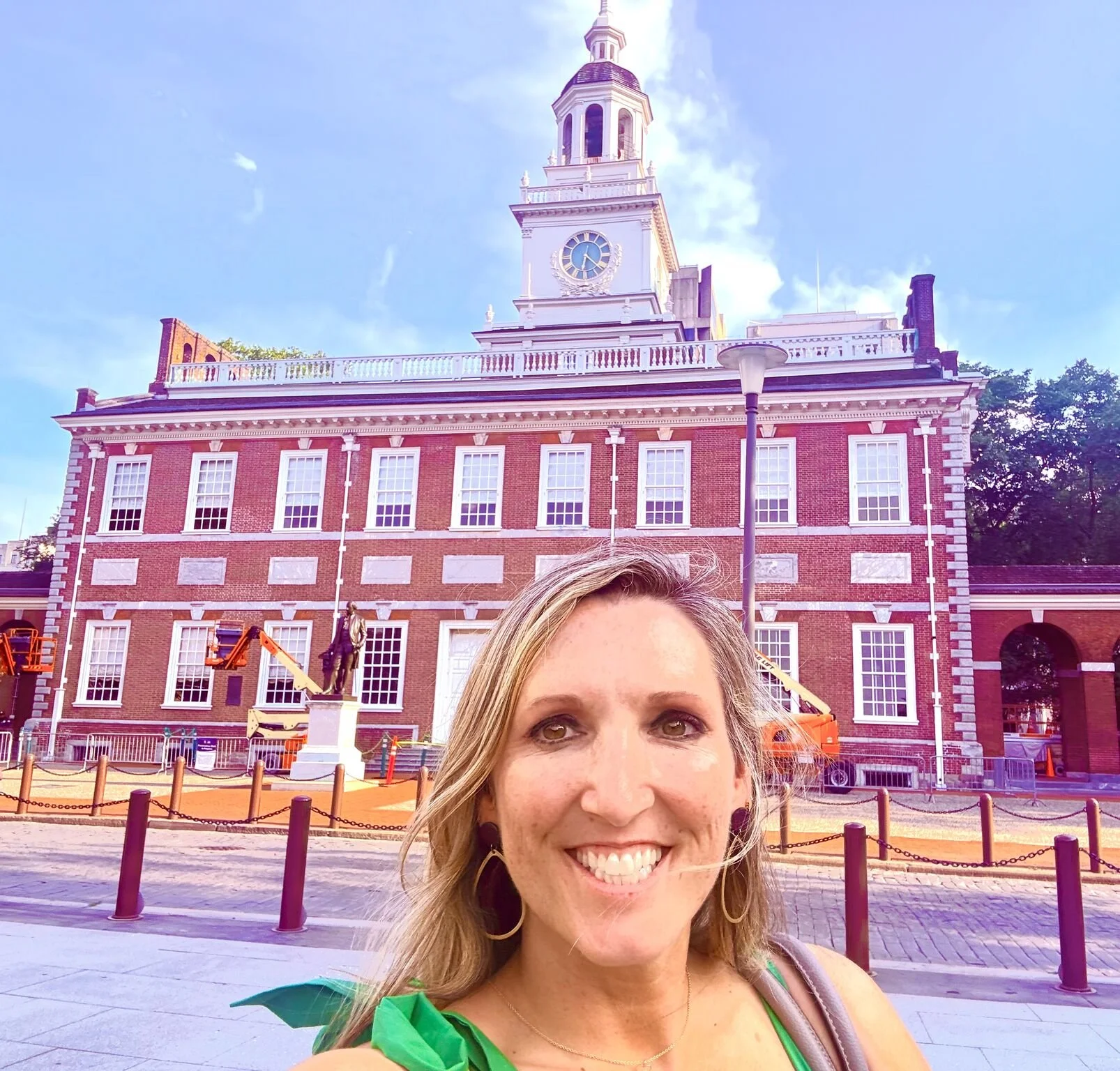 Jenny Hofler smiling takes a selfie in front of the Independence Hall in Philadelphia, Pennsylvania, with construction equipment nearby and a blue sky above.