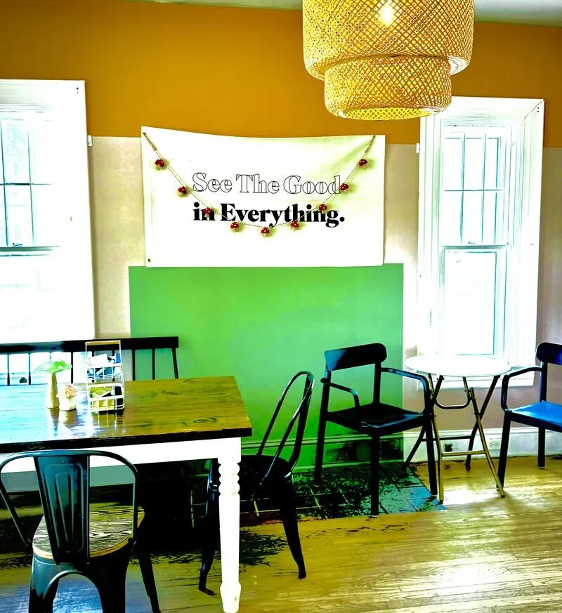 A cozy dining or sitting area with a wooden table, several black chairs, and a large window with white trim, with a white wall with an inspirational sign that reads 'See The Good in Everything' decorated with small red ornaments, and a large woven li