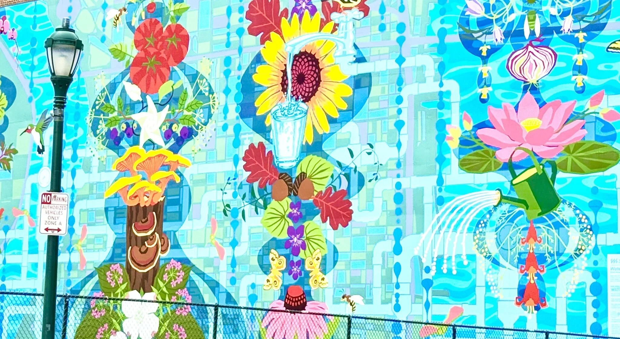 Colorful mural featuring flowers, greenery, hanging lanterns, hummingbirds, a watering can, and bees, with a blue patterned background.