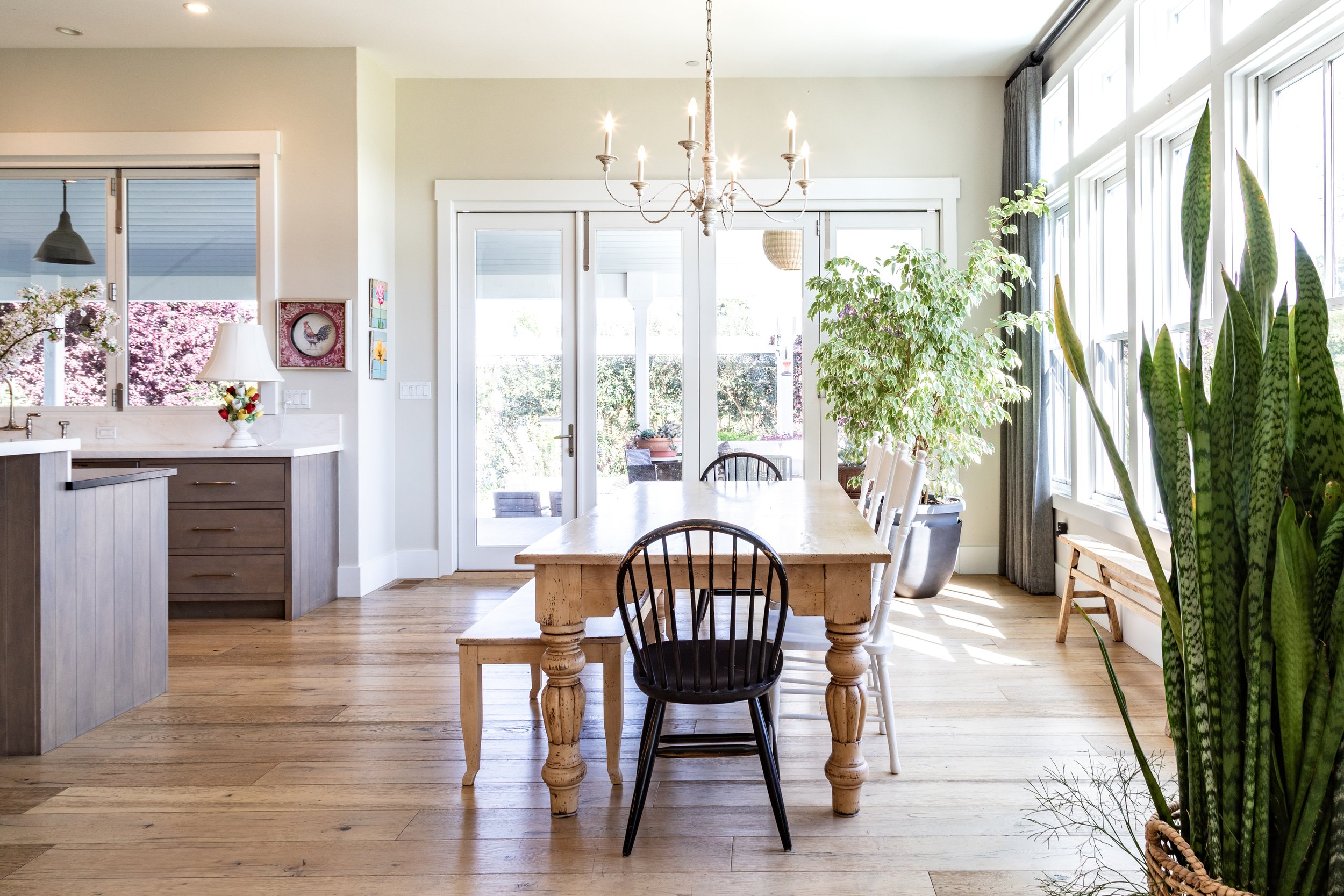 Bright dining room with a wooden table, black and white chairs, large windows with gray curtains, potted plants, a chandelier, and a sliding glass door leading outside.