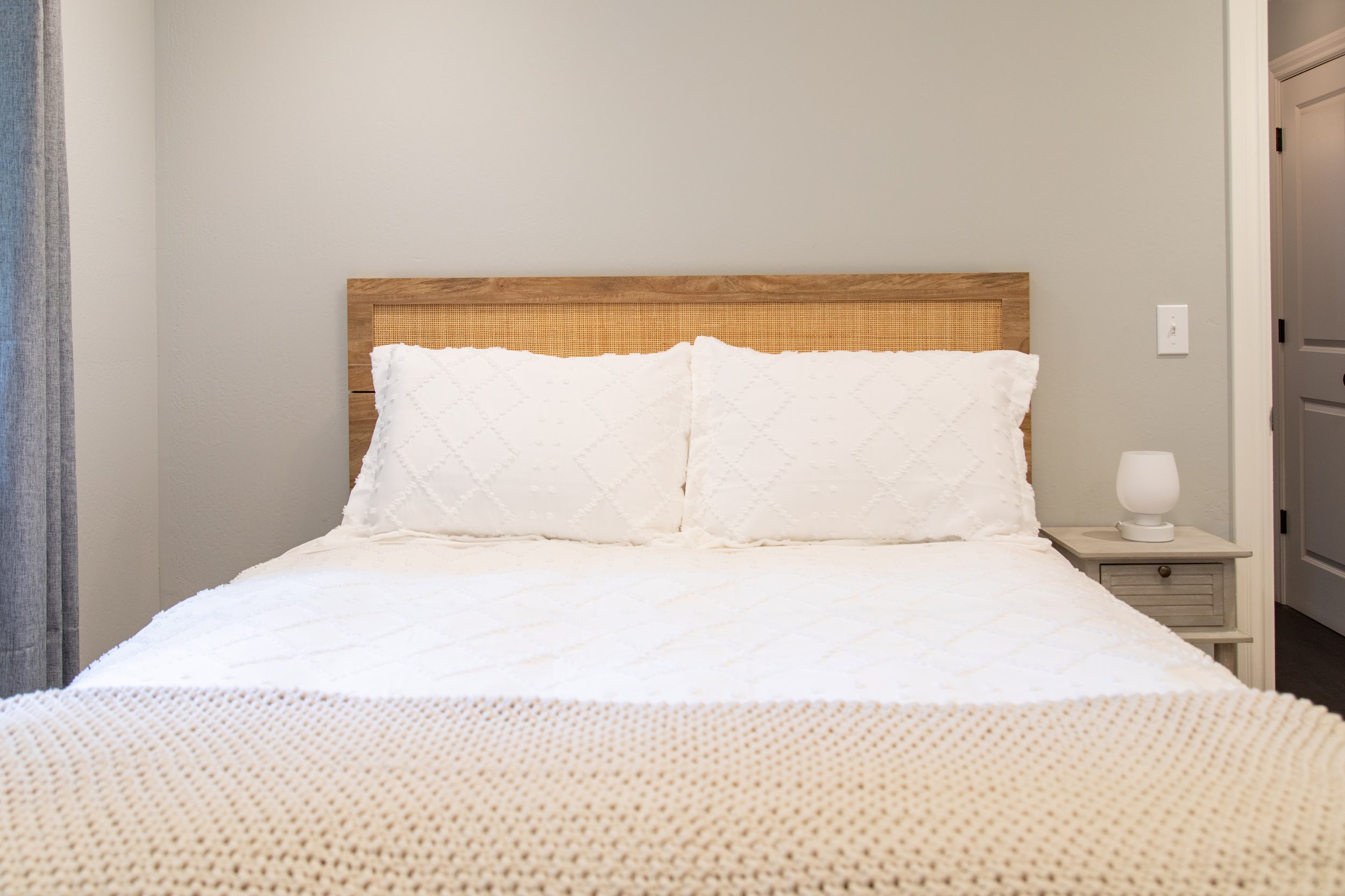 A bedroom with a bed covered in white bedding and two matching white pillows, a wooden headboard, a beige knitted blanket at the foot of the bed, a small white nightstand with a white lamp, and closed gray door on the right.