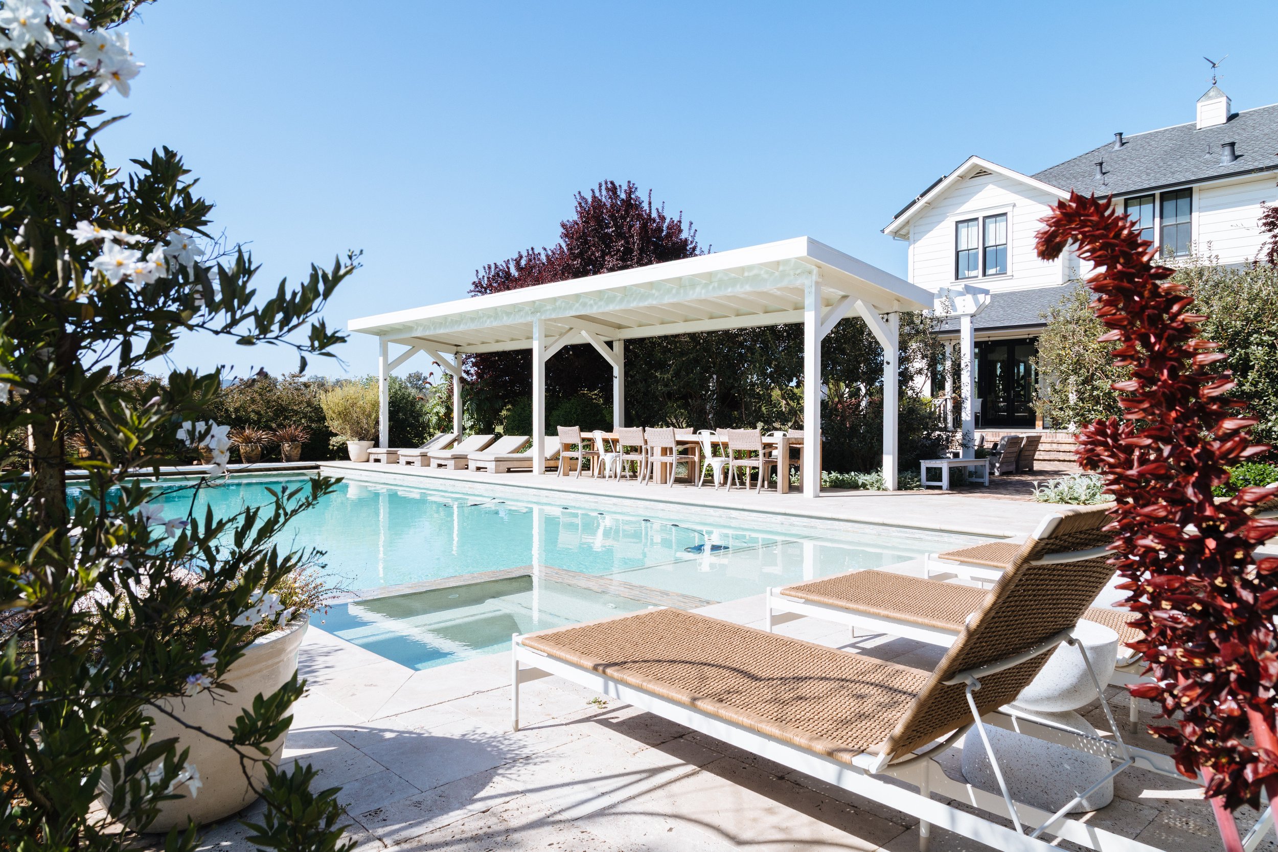 A backyard with a swimming pool, white lounge chairs, a white pergola, and a large white house with black window frames under a clear blue sky.